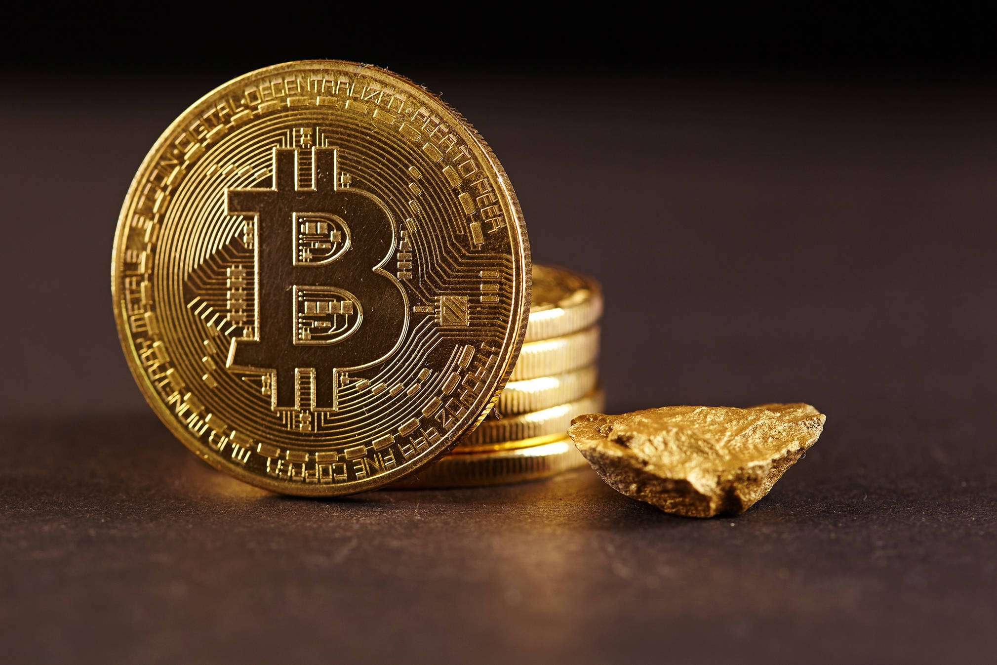 比特幣 桌布 壁紙 — 一枚閃閃發光的金色比特幣硬幣放在黑暗表面上的一塊原始金塊旁邊，象徵著數位和傳統財富。