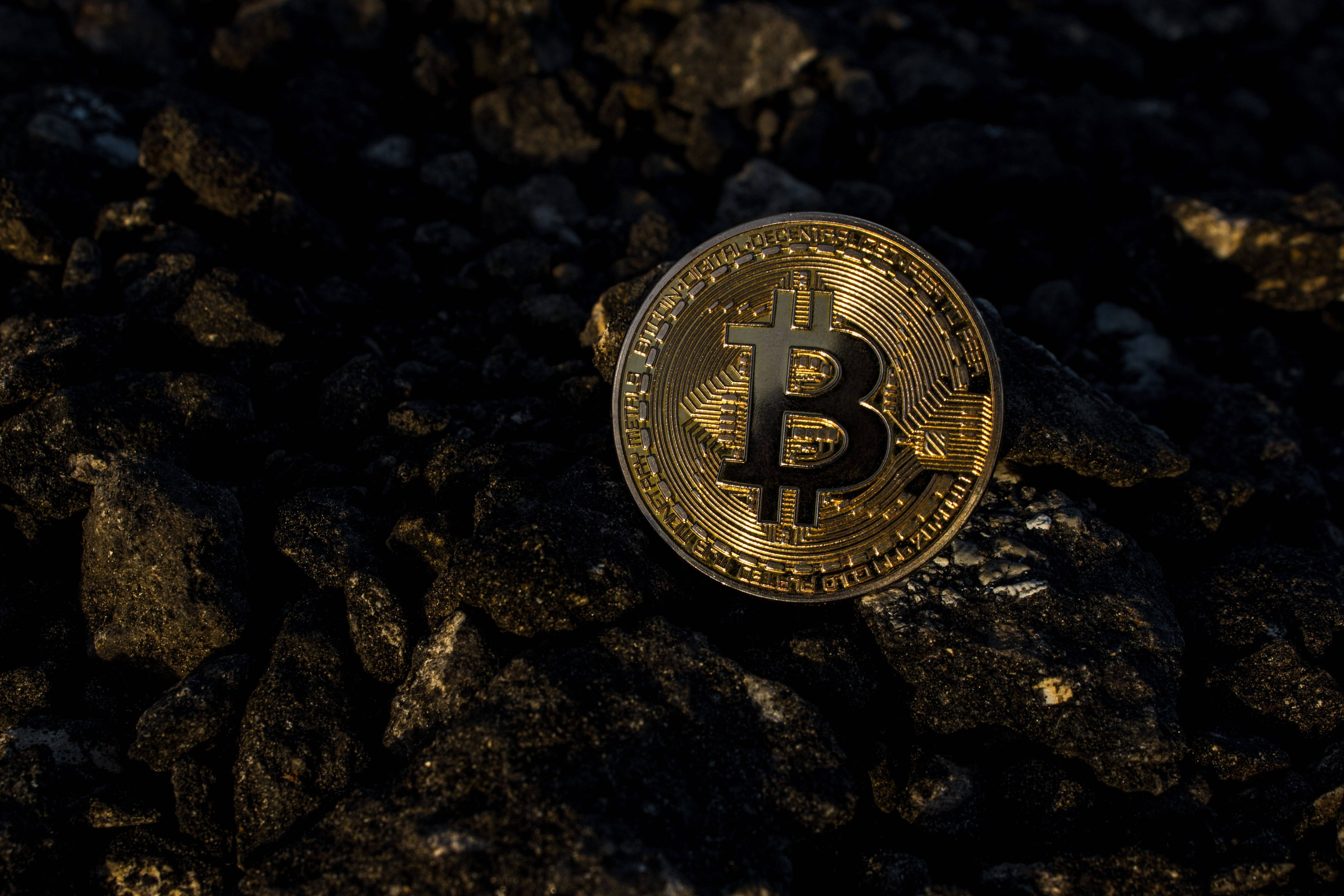 比特幣 桌布 壁紙 — 一枚閃閃發光的金色比特幣硬幣放在黑暗粗糙的岩石上，光線效果戲劇性。