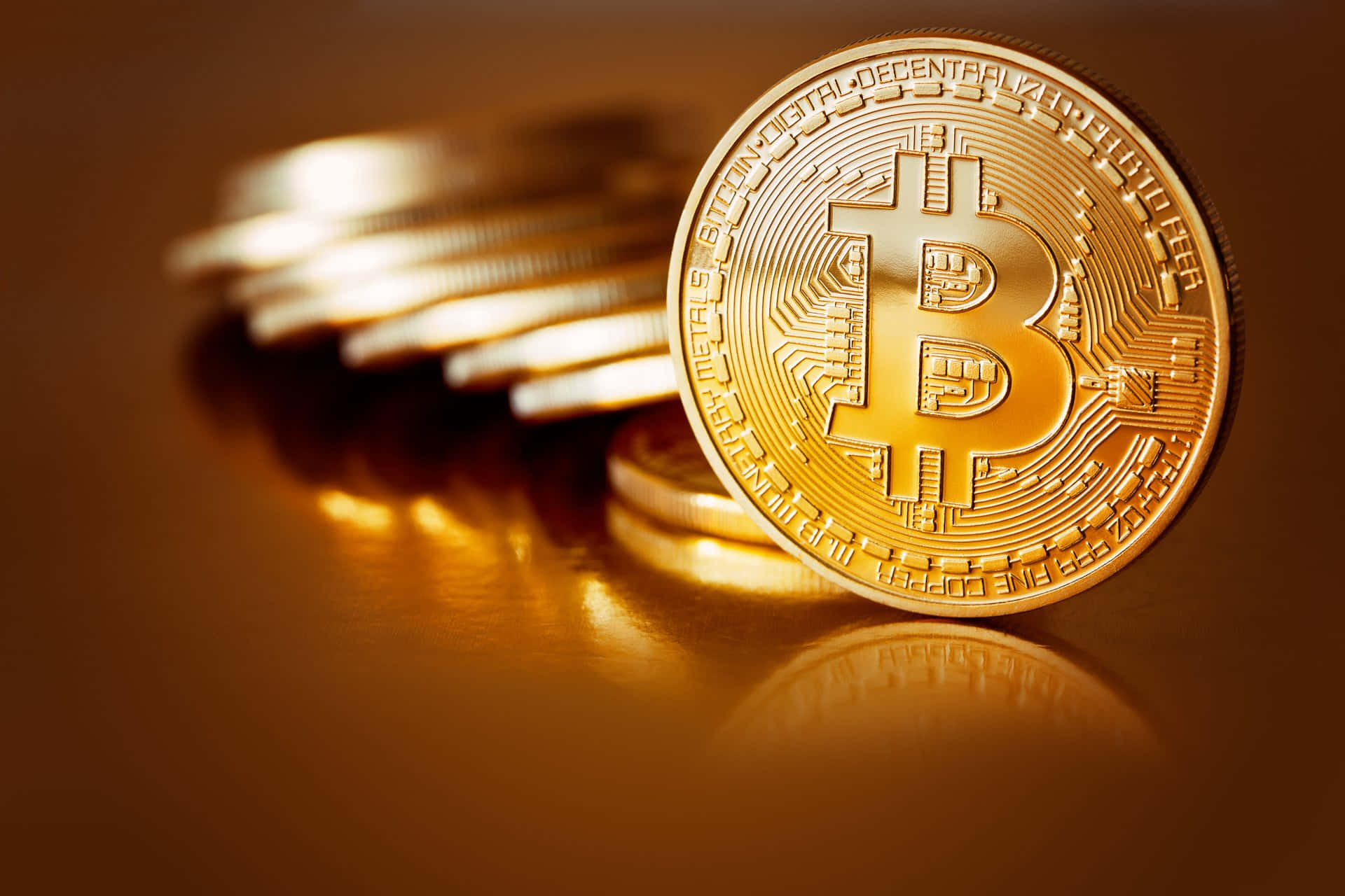 比特幣 桌布 壁紙 — 一枚閃閃發光的金色比特幣硬幣清晰對焦，旁邊是一堆模糊的同類硬幣，置於反光表面上。