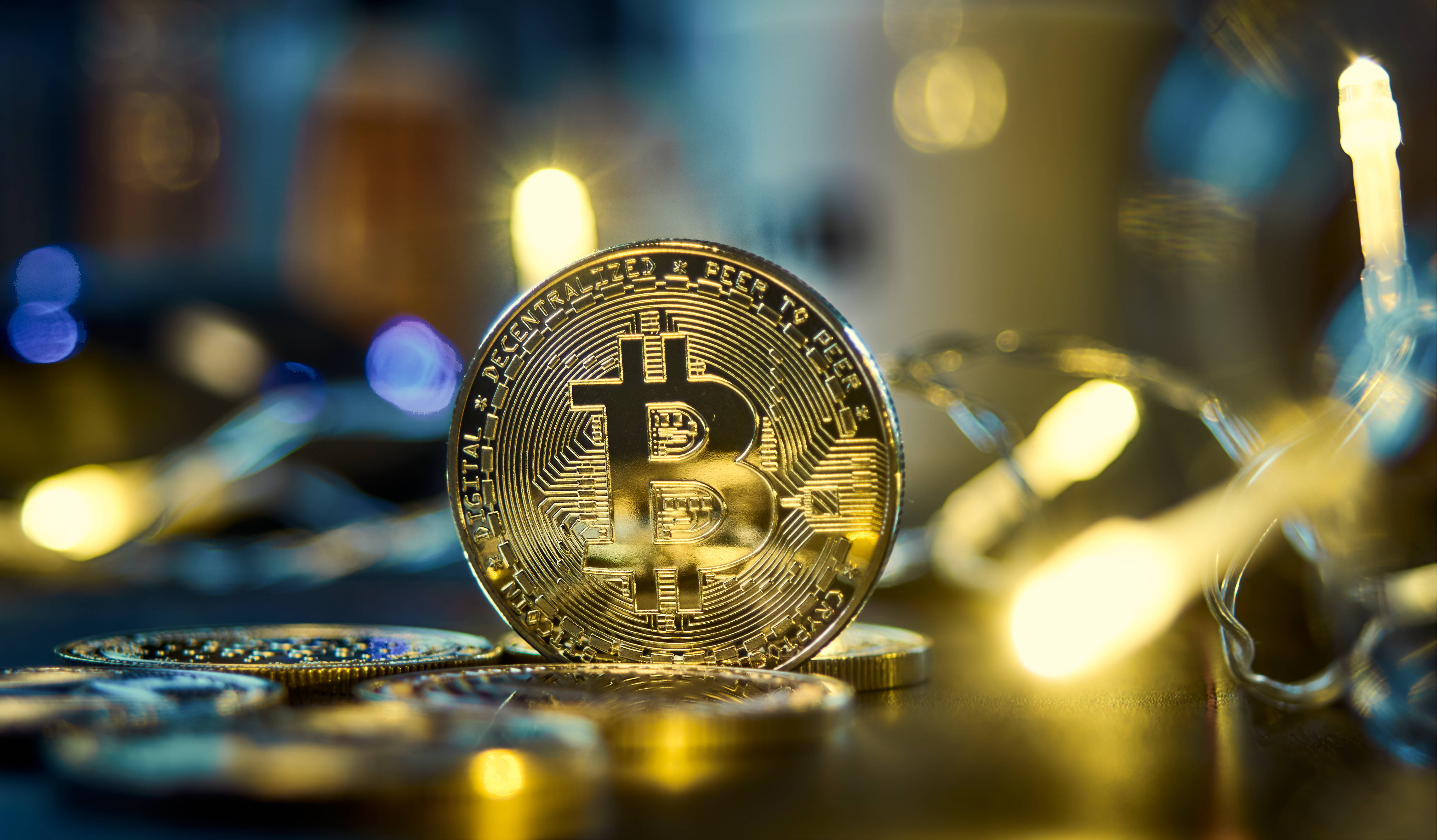 Cryptocurrency 桌布 壁紙 — 一枚閃閃發光的金色比特幣硬幣直立於散落的硬幣中，溫暖的仙女燈在模糊的背景中柔和地發光。