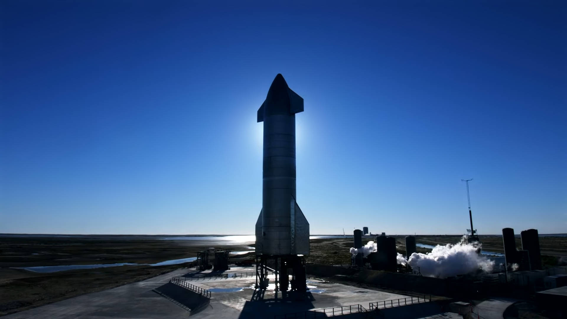 Spacex Starship 桌布 壁紙 — 一枚高聳的火箭在晴朗的藍天下呈現剪影，太陽在其後方照耀，附近的設備冒出蒸氣。