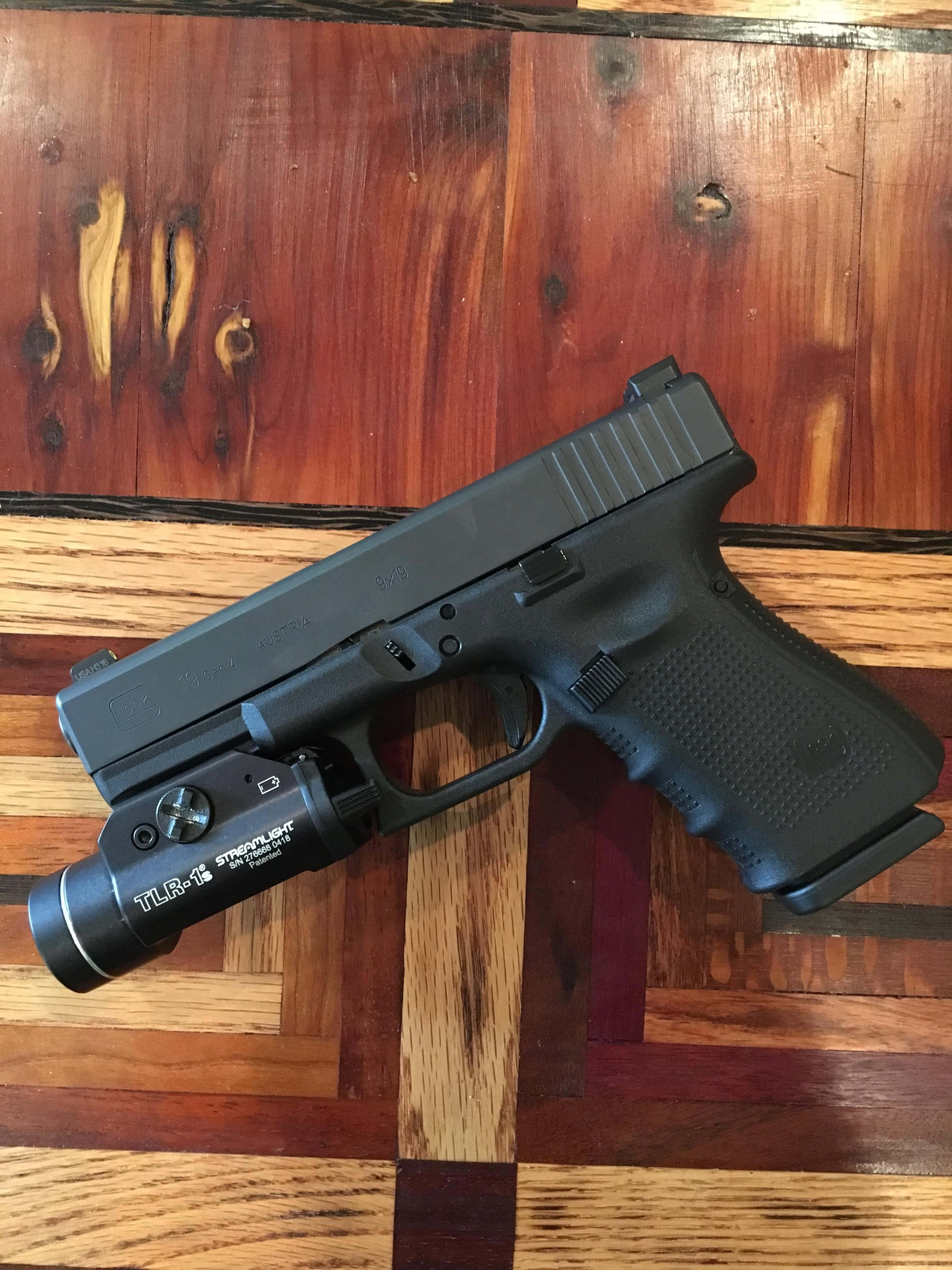 Glock 19 桌布 壁紙 — 一枝黑色手槍連戰術燈放在一塊紋理豐富、帶有天然木節和不同色調的木質表面上。