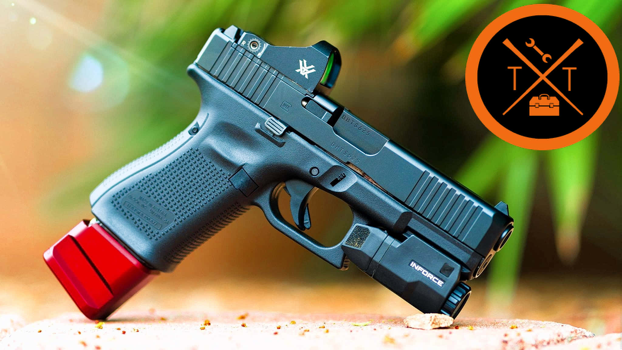 Glock 19 桌布 壁紙 — 一枝黑色手槍配紅色彈匣及戰術燈，擱在沙地上，背景是模糊的綠色樹葉。