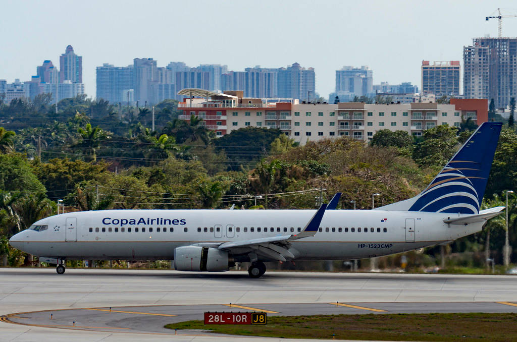 飛機 桌布 壁紙 — 一架 Copa Airlines 的波音 737 在陽光明媚的機場停機坪上滑行，背景是密集的城市天際線和茂密的綠色植物。