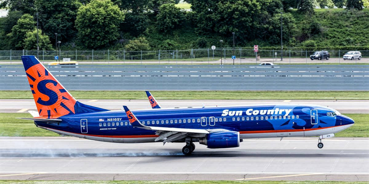 Airport 桌布 壁紙 — 一架 Sun Country Airlines 的波音 737 在跑道上滑行，背景有茂密的綠樹和遠處的汽車。