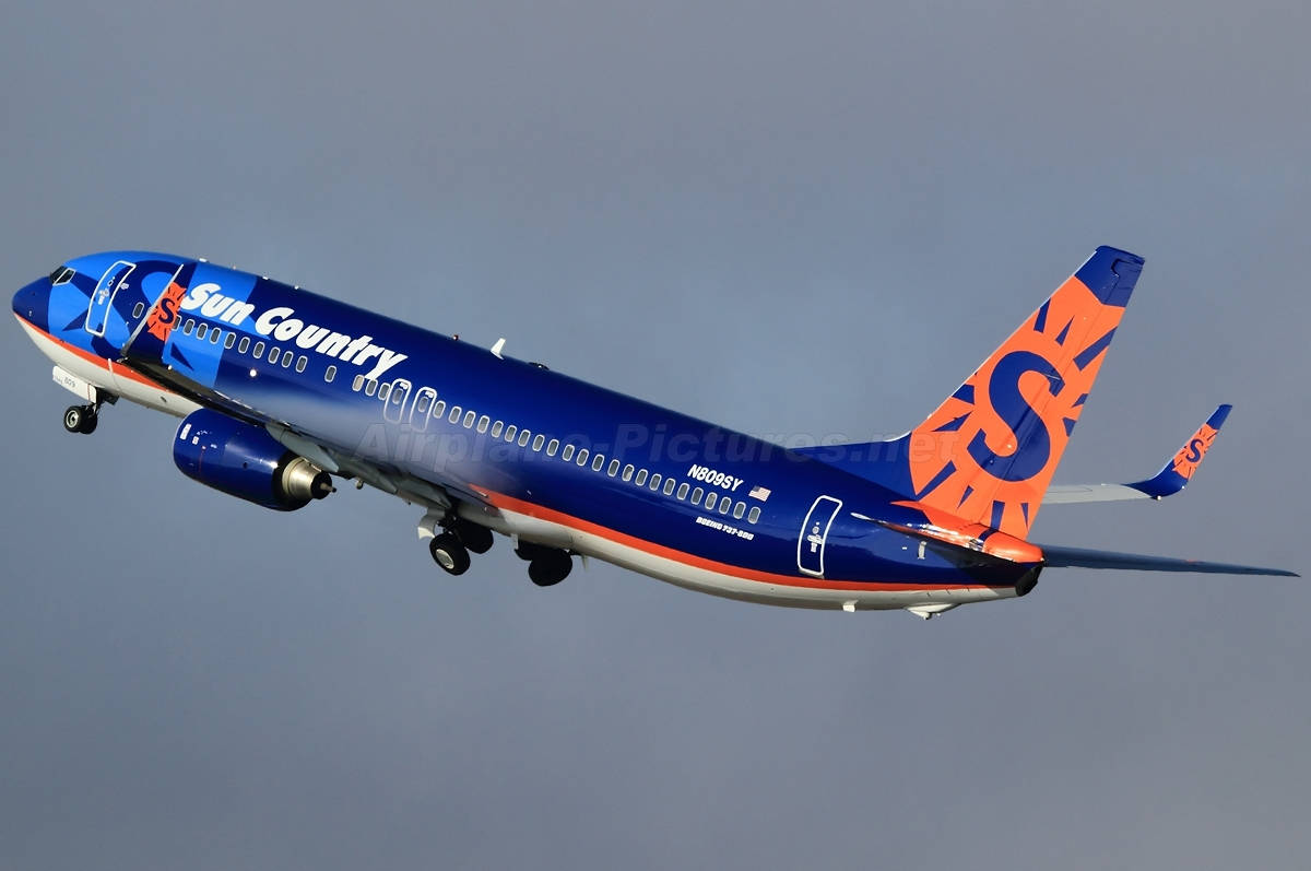 飛機 桌布 壁紙 — 一架 Sun Country Airlines 的波音 737 以鮮豔的藍色和橙色塗裝，在晴朗的灰色天空中翱翔，起落架已放下。