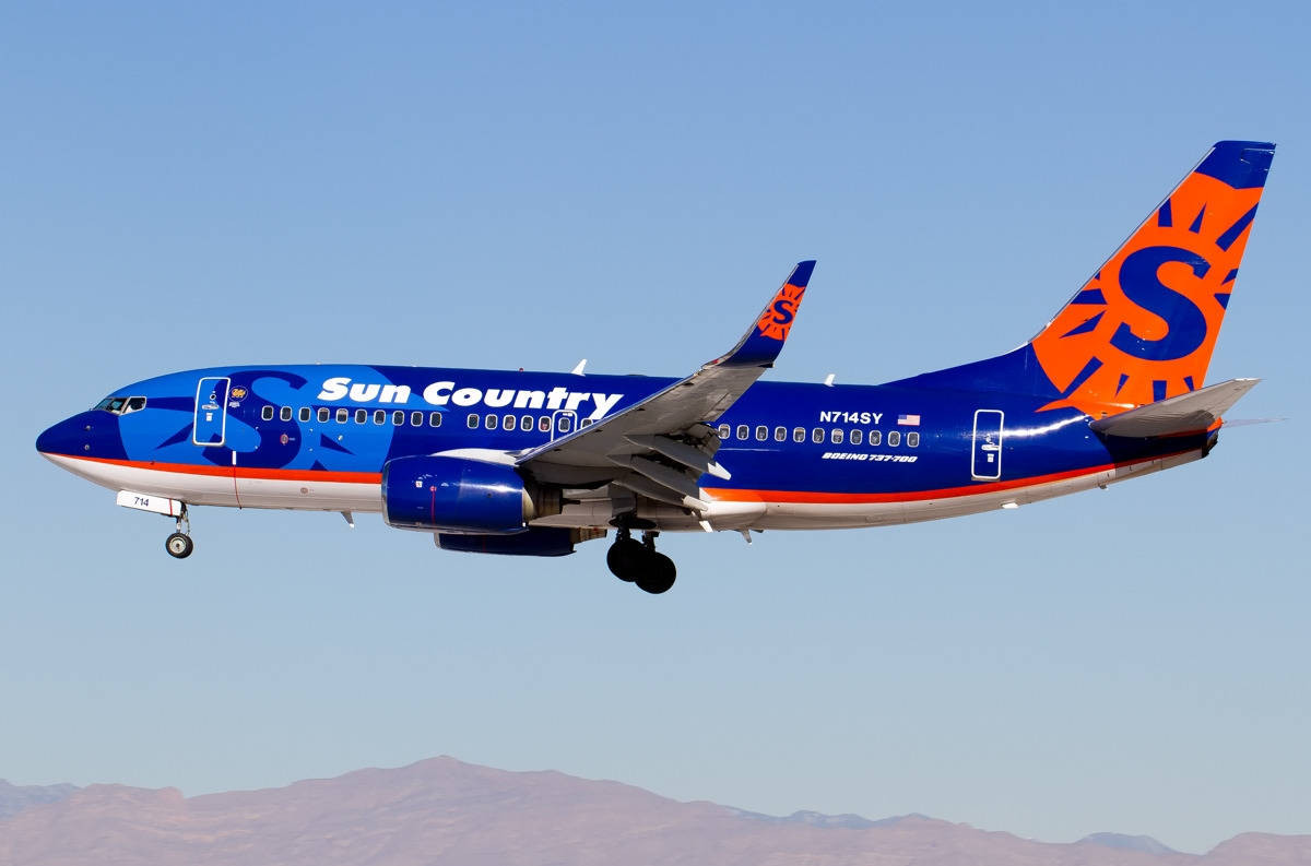 Aviation 桌布 壁紙 — 一架 Sun Country Airlines 的波音 737 以鮮豔的藍色和橙色塗裝，在晴朗的藍天中翱翔，下方是遠山。