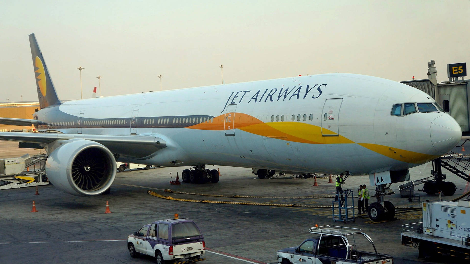  壁紙 — 一架Jet Airways波音777停泊在機場閘口，附近有地勤人員和服務車輛，天空帶有薄霧。