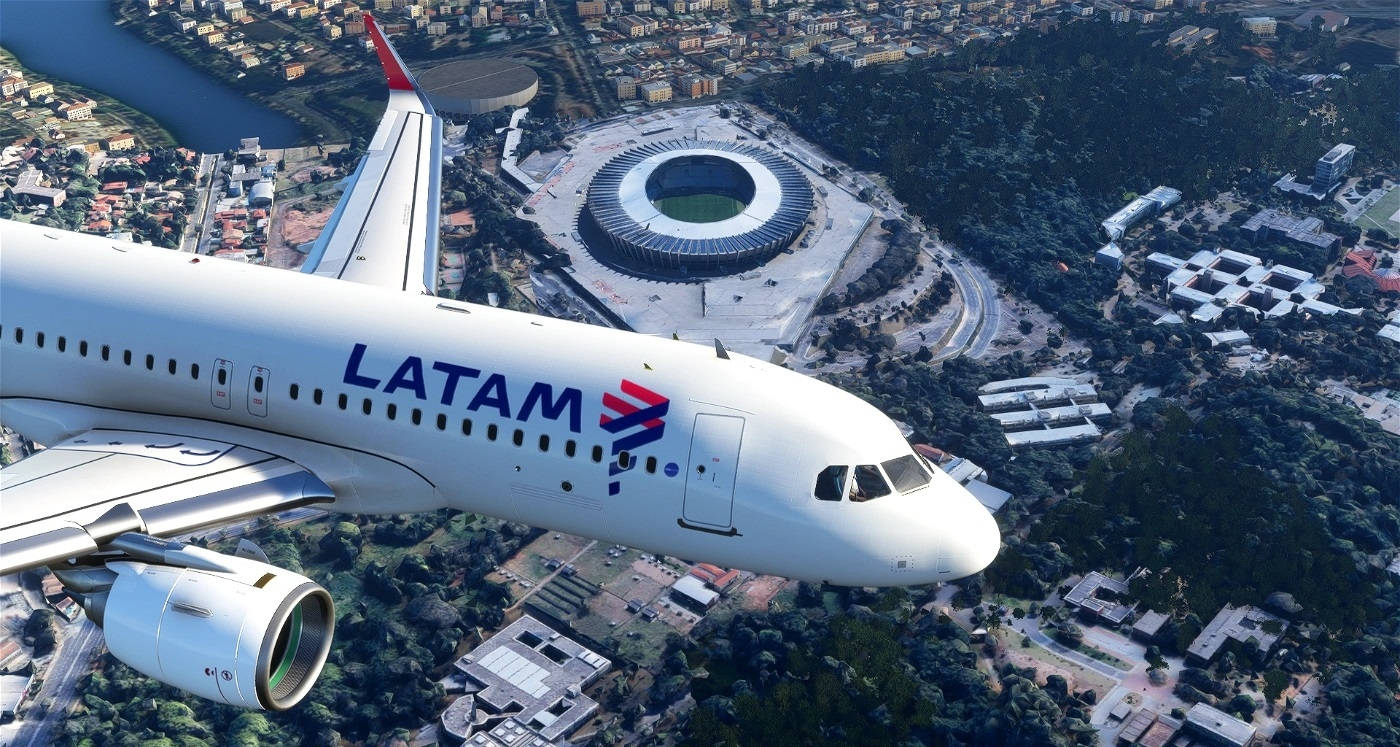 flying 桌布 壁紙 — 一架LATAM飛機翱翔於廣闊的城市景觀之上，其中有一個突出的圓形體育館坐落在綠丘和城市社區之間。
