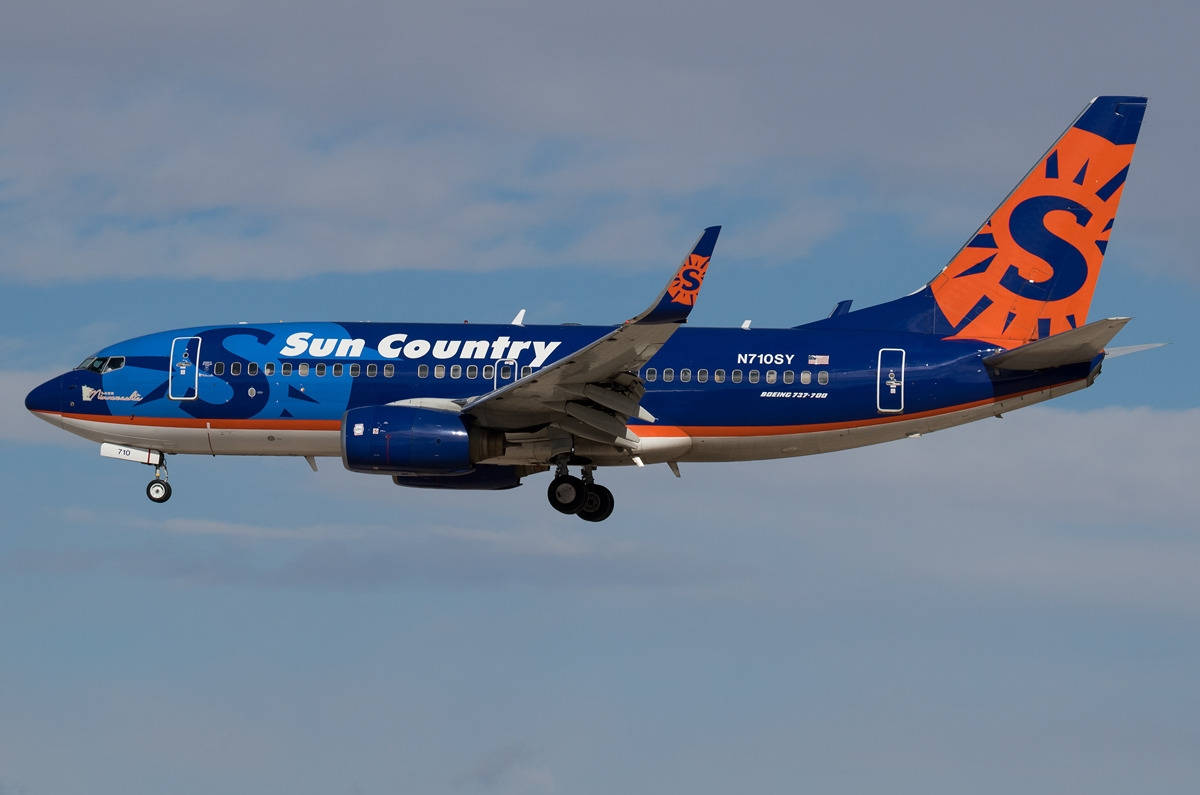  壁紙 — 一架Sun Country Airlines的波音737，藍橙塗裝，起落架放下，傾斜飛過多雲的天空。