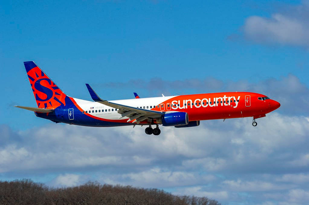 Aviation 桌布 壁紙 — 一架Sun Country Airlines的噴射機，以紅、藍、白塗裝，在部分多雲的天空和下方的光禿樹木間傾斜飛行。