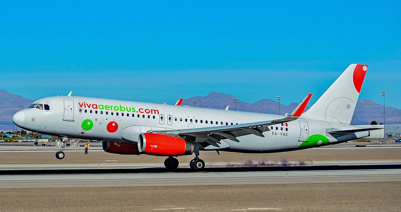 Airport 桌布 壁紙 — 一架Viva Aerobus飛機，紅色引擎，綠色紅色圓形標誌，在晴朗藍天和遠山背景下在跑道上滑行。