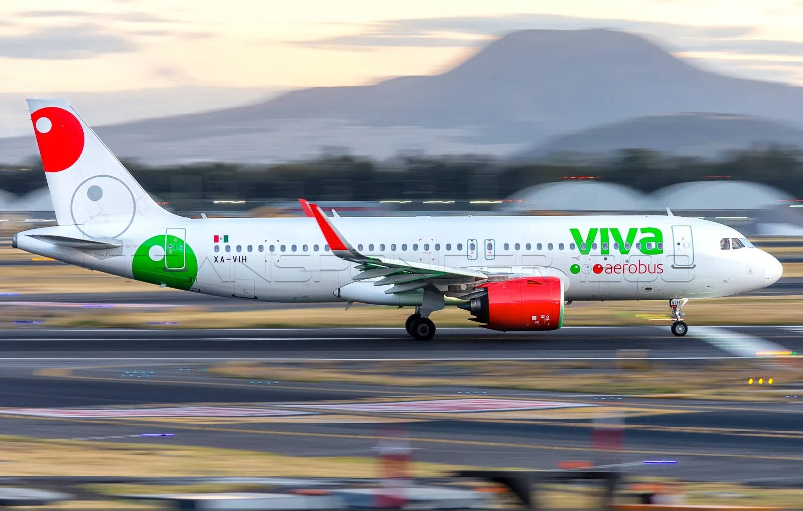 Airport 桌布 壁紙 — 一架Viva Aerobus飛機在黃昏時分在跑道上加速，帶有模糊的動態和遠處山脈在柔和粉彩光中。