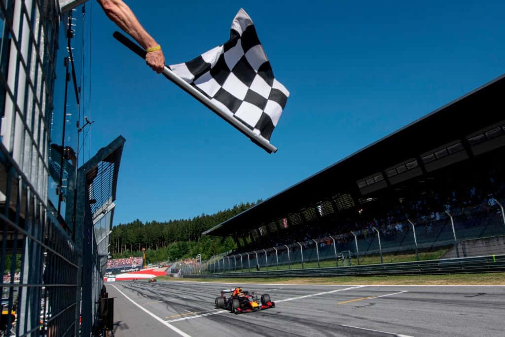 Chequered Flag 桌布 壁紙 — 一架一級方程式賽車越過終點線，同時有一隻手在晴朗的藍天下揮動方格旗。