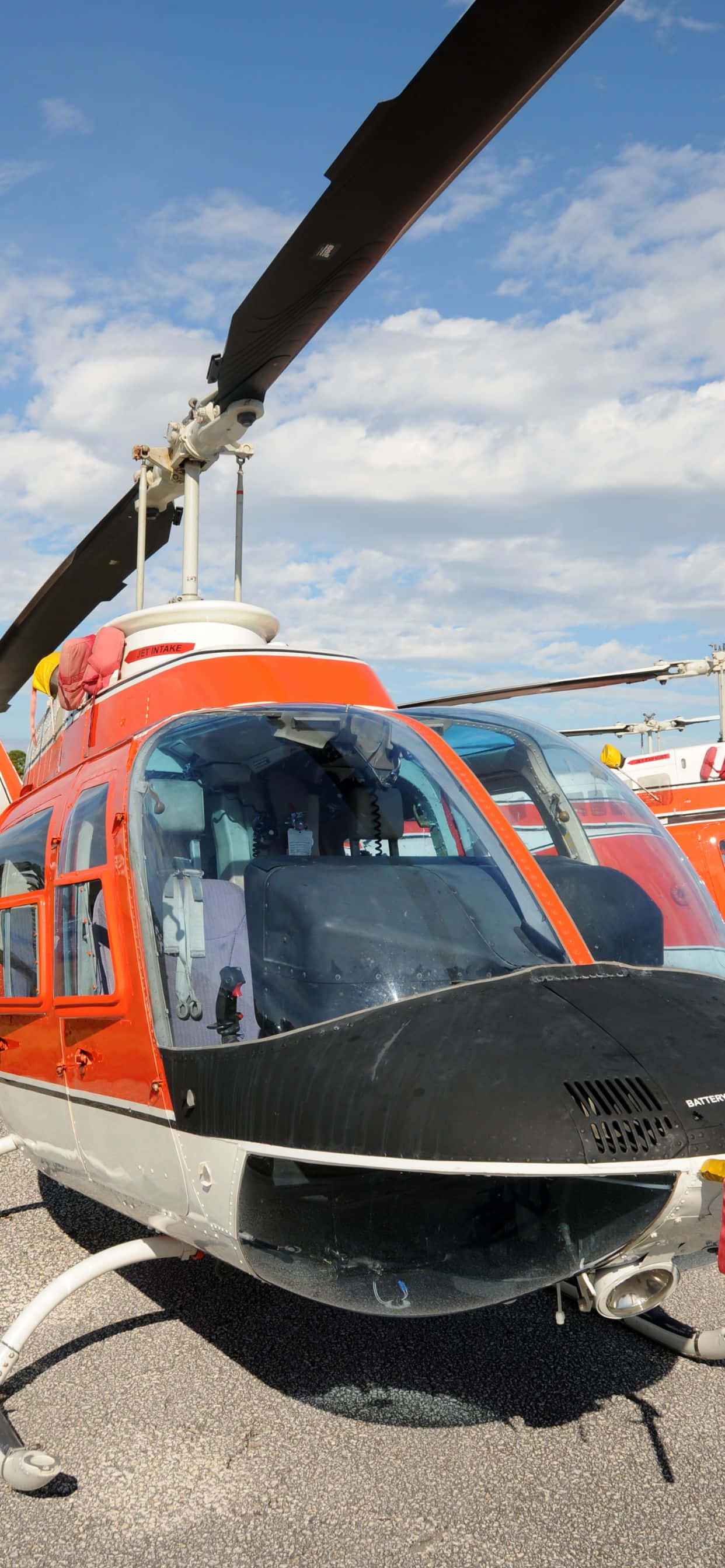iPhone Xs Max Helicopters 桌布 壁紙 — 一架亮橙色和黑色的直升機停放在停機坪上，天空部分多雲，呈現藍色。