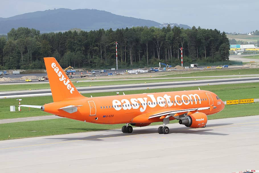 Airport 桌布 壁紙 — 一架亮橙色的 easyJet 飛機在跑道上滑行，背景是綠色田野和森林覆蓋的山丘，天空多雲。
