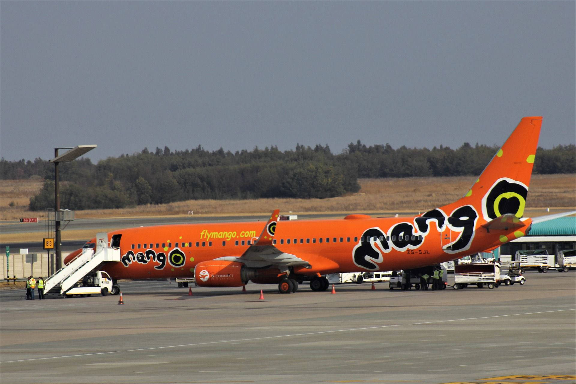 飛機 桌布 壁紙 — 一架亮橙色的Mango Airlines飛機停在機場閘口，附有登機梯，附近有地勤人員。