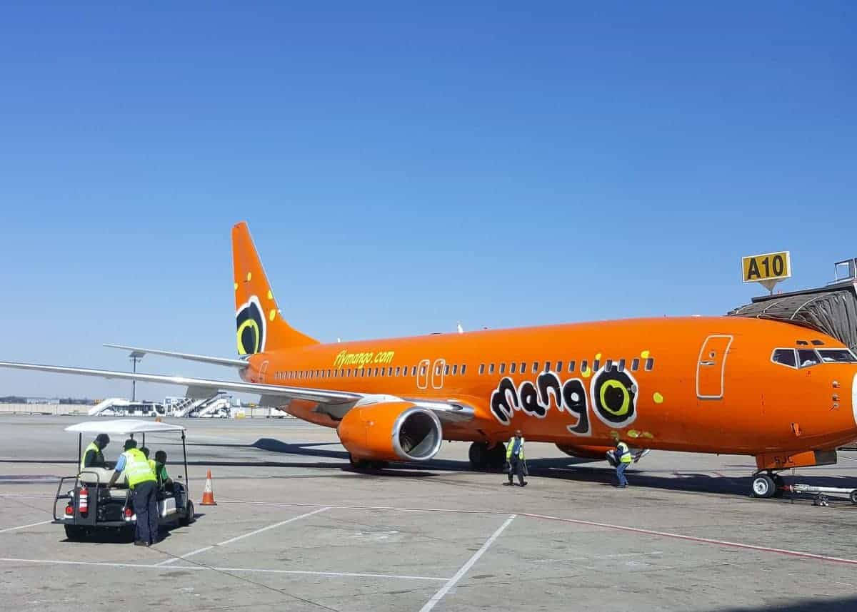 Airport 桌布 壁紙 — 一架亮橙色的Mango Airlines飛機停泊在A10登機門，附近有地勤人員和手推車，在晴朗藍天下。