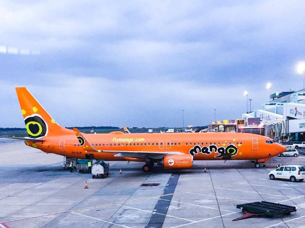 飛機 桌布 壁紙 — 一架亮橙色的Mango Airlines飛機在陰雲密佈的黃昏天空下停泊在機場閘口。
