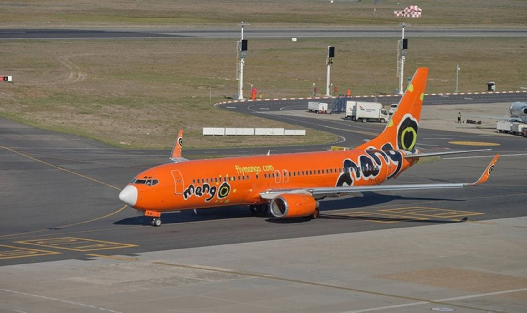 飛機 桌布 壁紙 — 一架亮橙色的Mango Airlines飛機在陽光明媚的機場停機坪上滑行，跑道標記和地面設備清晰可見。