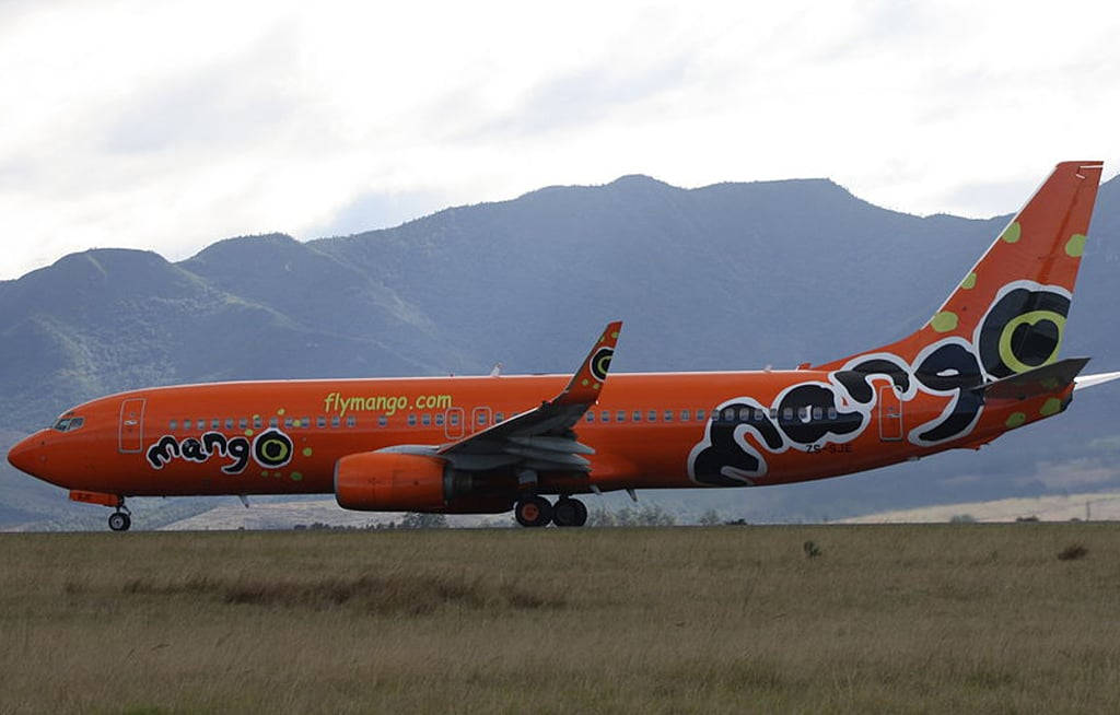 山景 桌布 壁紙 — 一架亮橙色的Mango Airlines飛機在草地上滑行，背景是綠色的山脈，天空多雲。