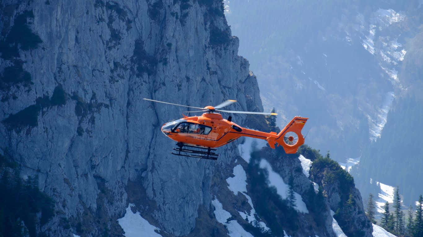 1366x768 Helicopters 桌布 壁紙 — 一架亮橙色的救援直升機低飛，靠近一座高聳、積雪覆蓋的山崖面。