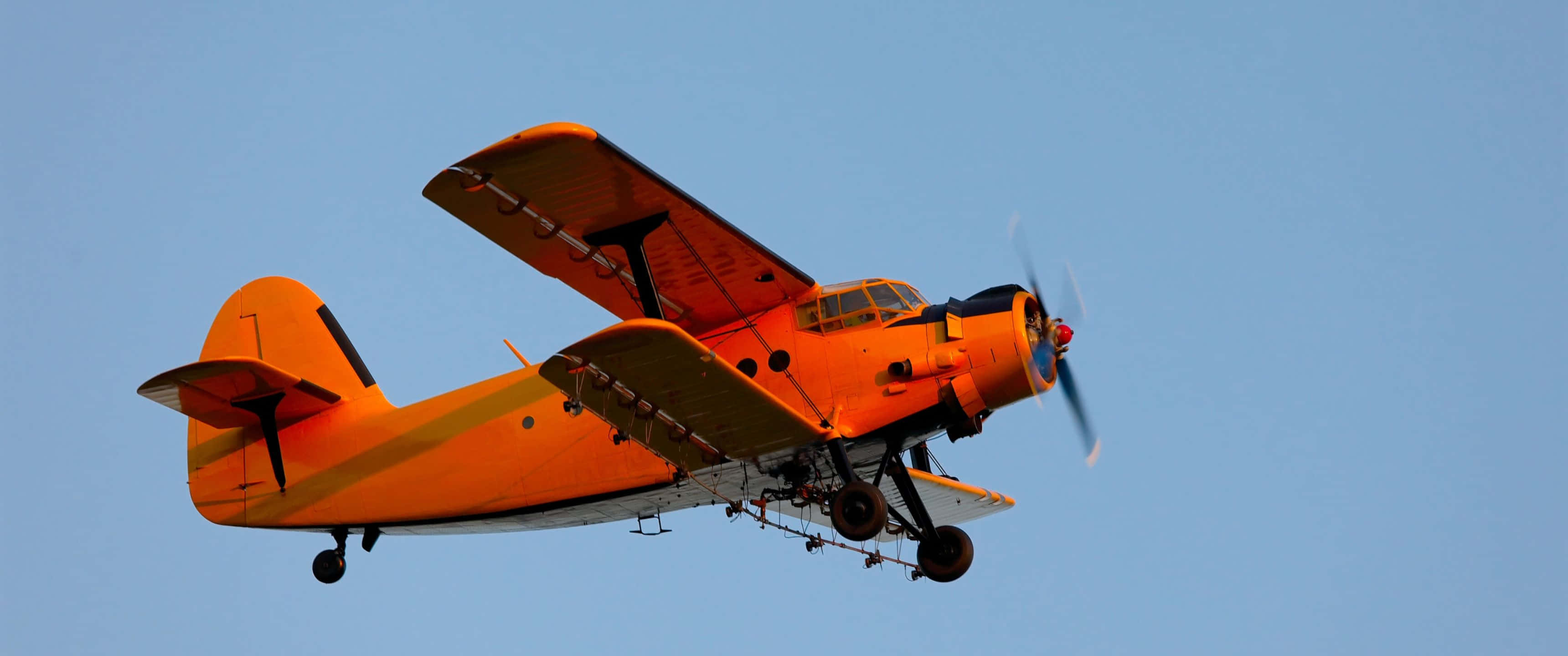 3440x1440p Small Planes 桌布 壁紙 — 一架亮橙色的消防飛機在清澈的藍天中翱翔，螺旋槳旋轉，投放箱展開。