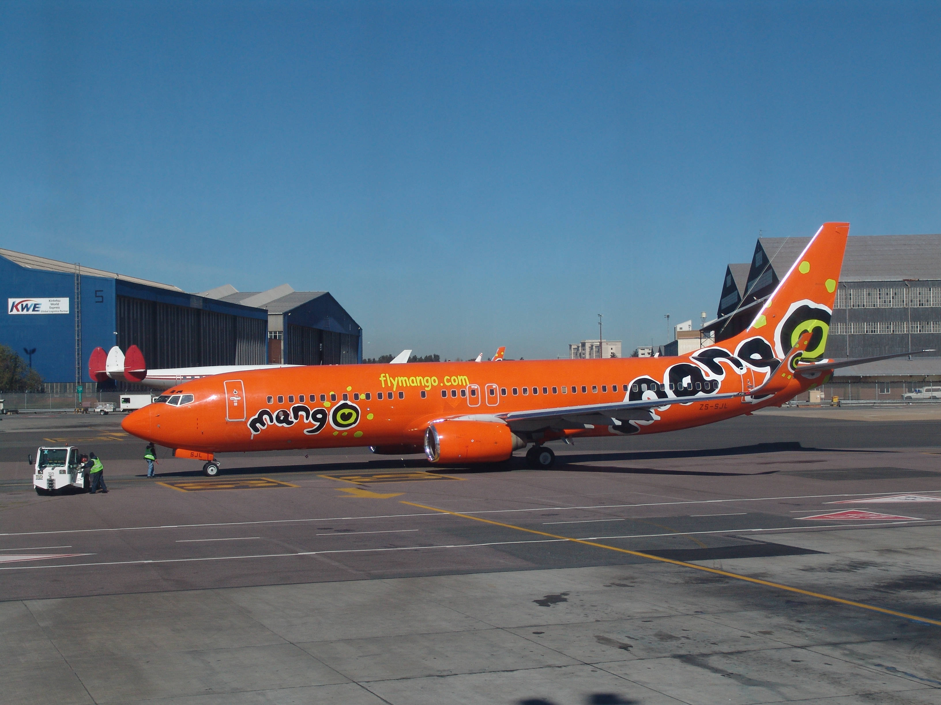 飛機 桌布 壁紙 — 一架亮橙色的芒果航空飛機停在機場停機坪上，天空湛藍，附近有地勤人員。