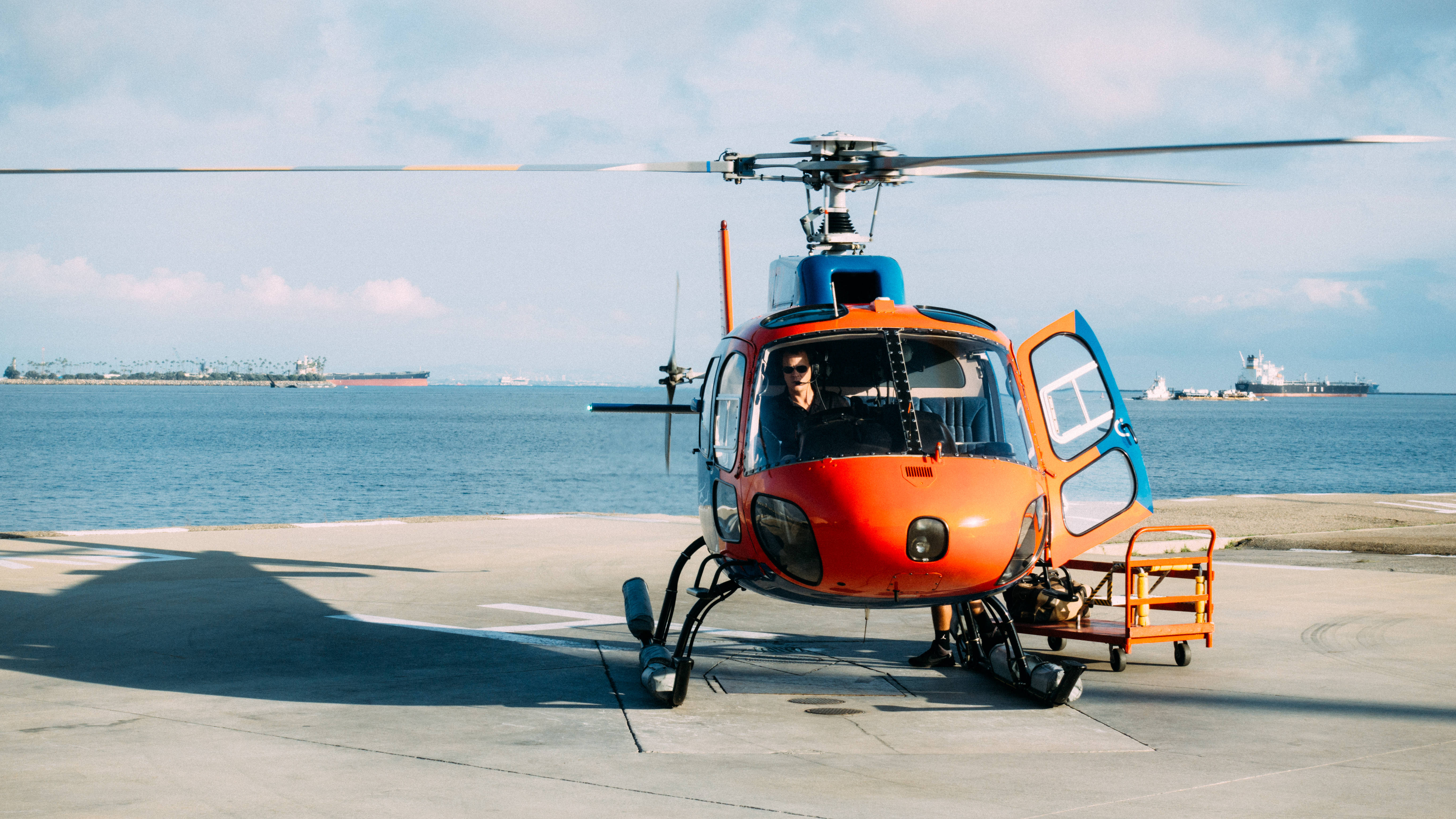 helicopter 4K 桌布 壁紙 — 一架亮橙色直升機停在海邊的混凝土停機坪上，遠處可見船隻，天空部分多雲。