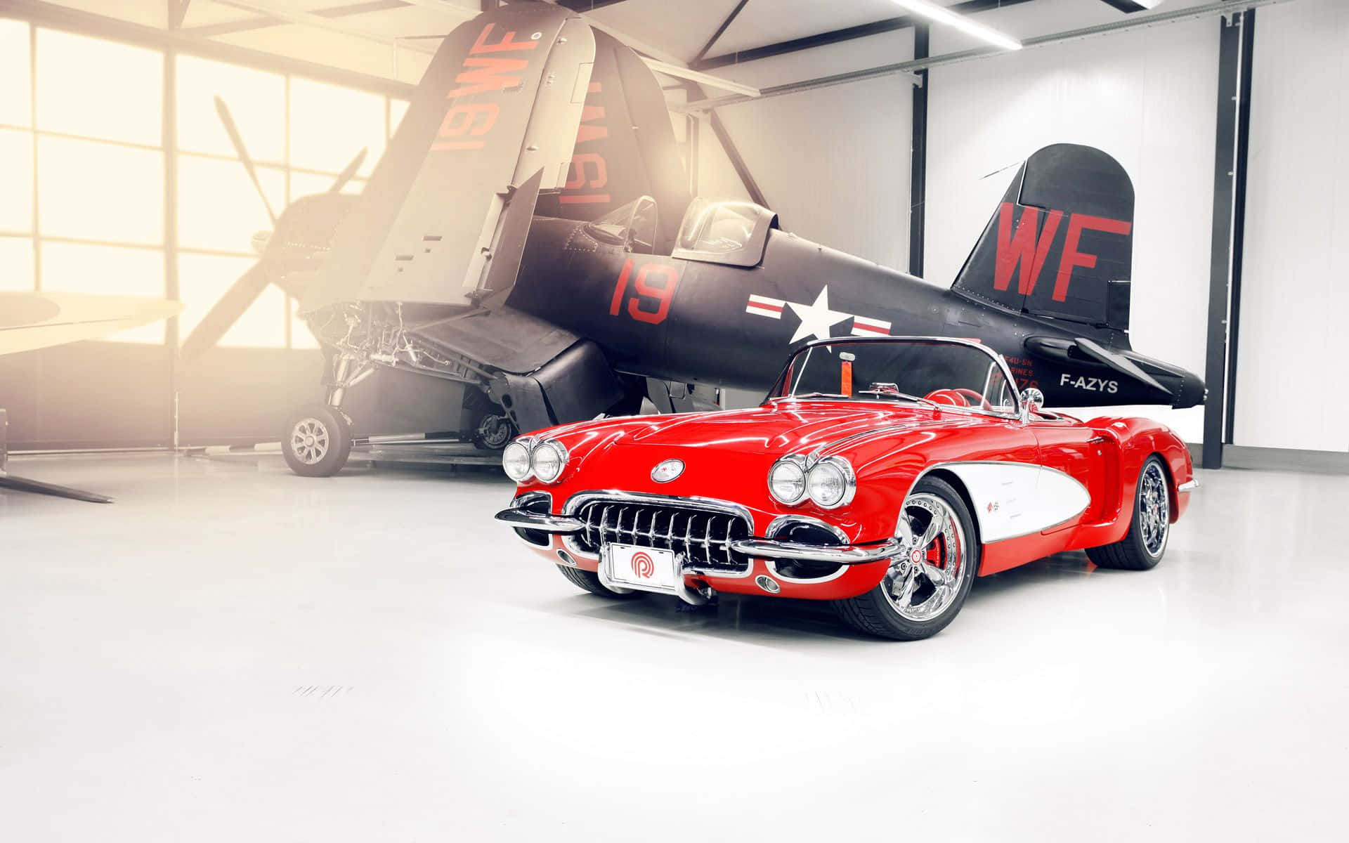 classic 桌布 壁紙 — 一架亮面紅色經典Corvette敞篷車停放在明亮的機庫內，前面是一架復古軍用戰鬥機。