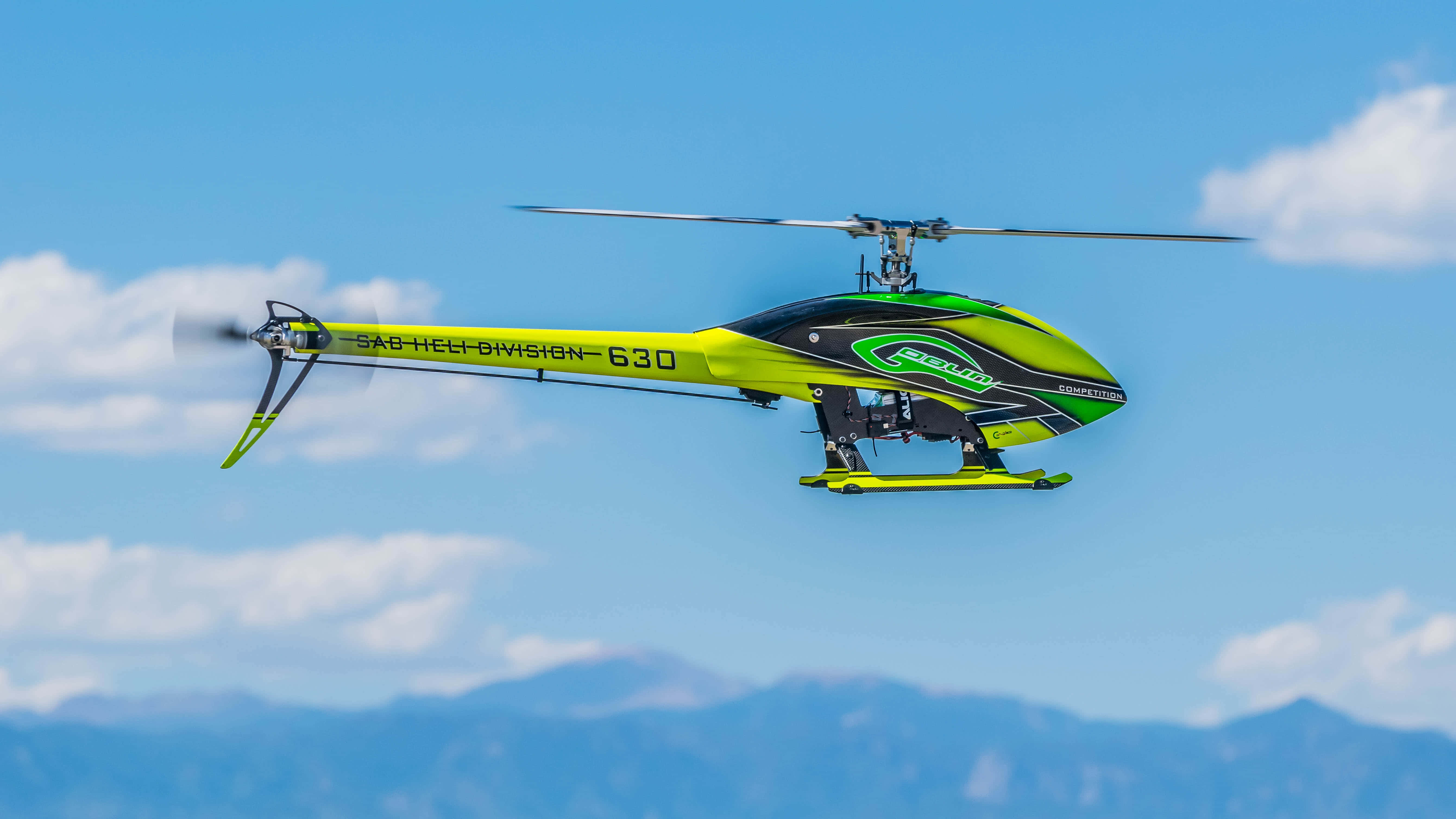 Helicopter 桌布 壁紙 — 一架亮黃色和綠色的遙控直升機在晴朗的藍天中翱翔，下方是遙遠的山脈。