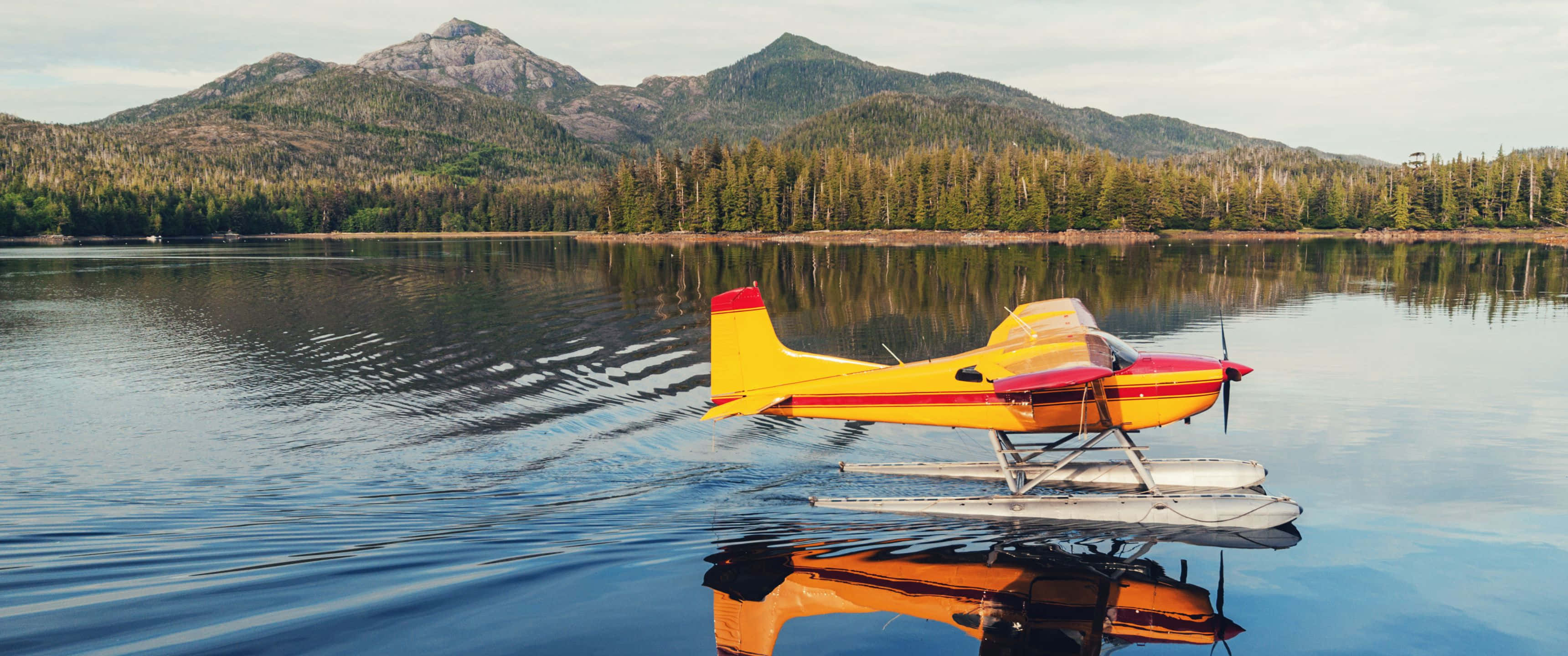 3440x1440p Small Planes 桌布 壁紙 — 一架亮黃色水上飛機帶有紅色點綴，平靜地漂浮在玻璃般的湖面上，四周是森林覆蓋的山脈，天空柔和。