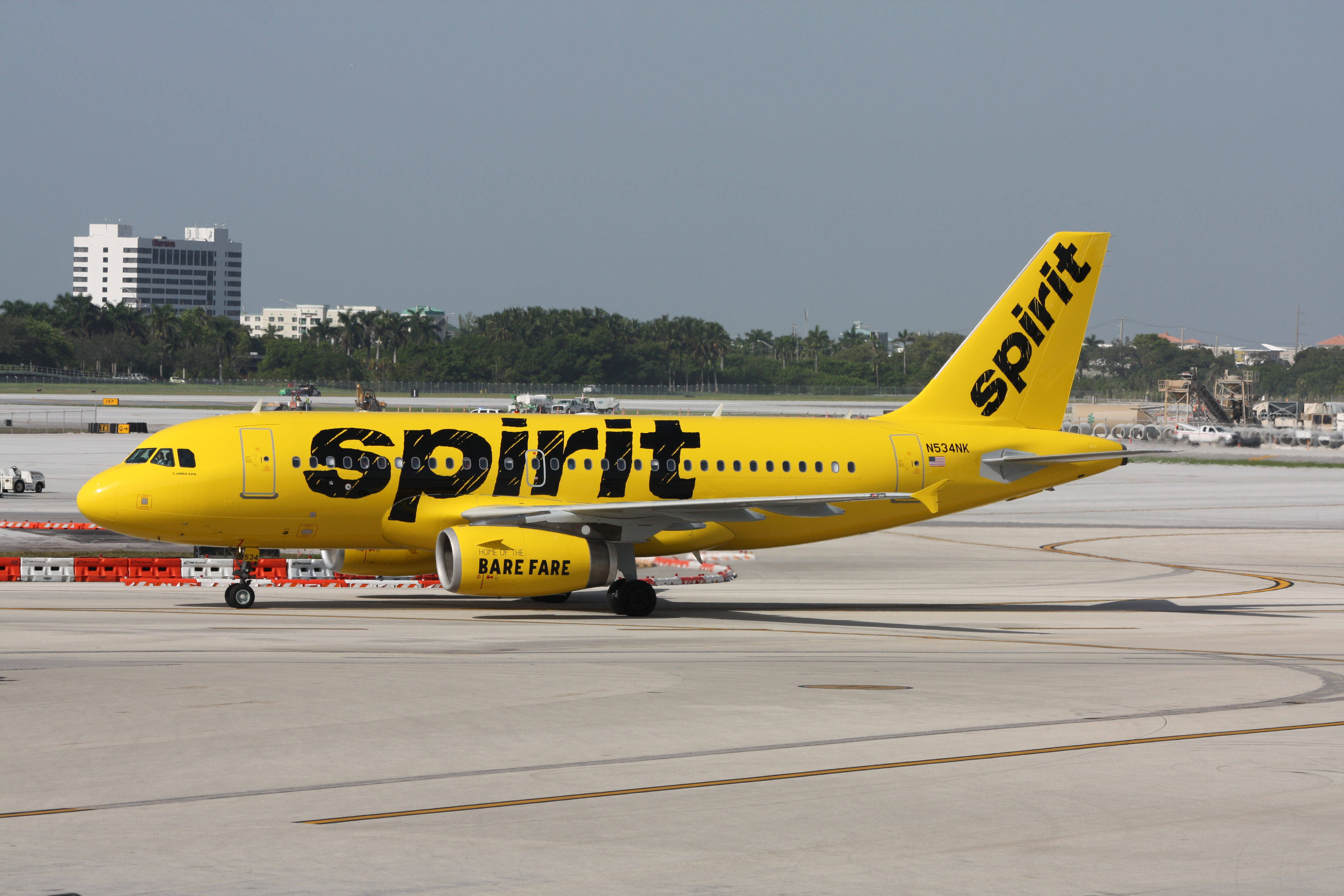 飛機 桌布 壁紙 — 一架亮黃色的Spirit Airlines飛機帶有「bare fare」品牌，在陽光明媚的機場停機坪上滑行，背景有棕櫚樹和建築物。