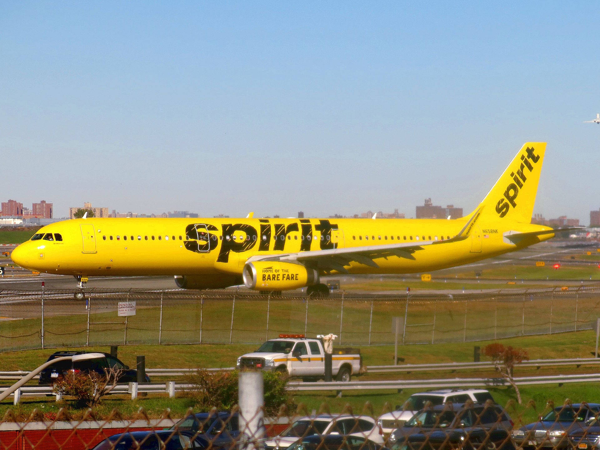  壁紙 — 一架亮黃色的Spirit Airlines噴射機在跑道上滑行，背景是城市天際線，天空清澈湛藍。