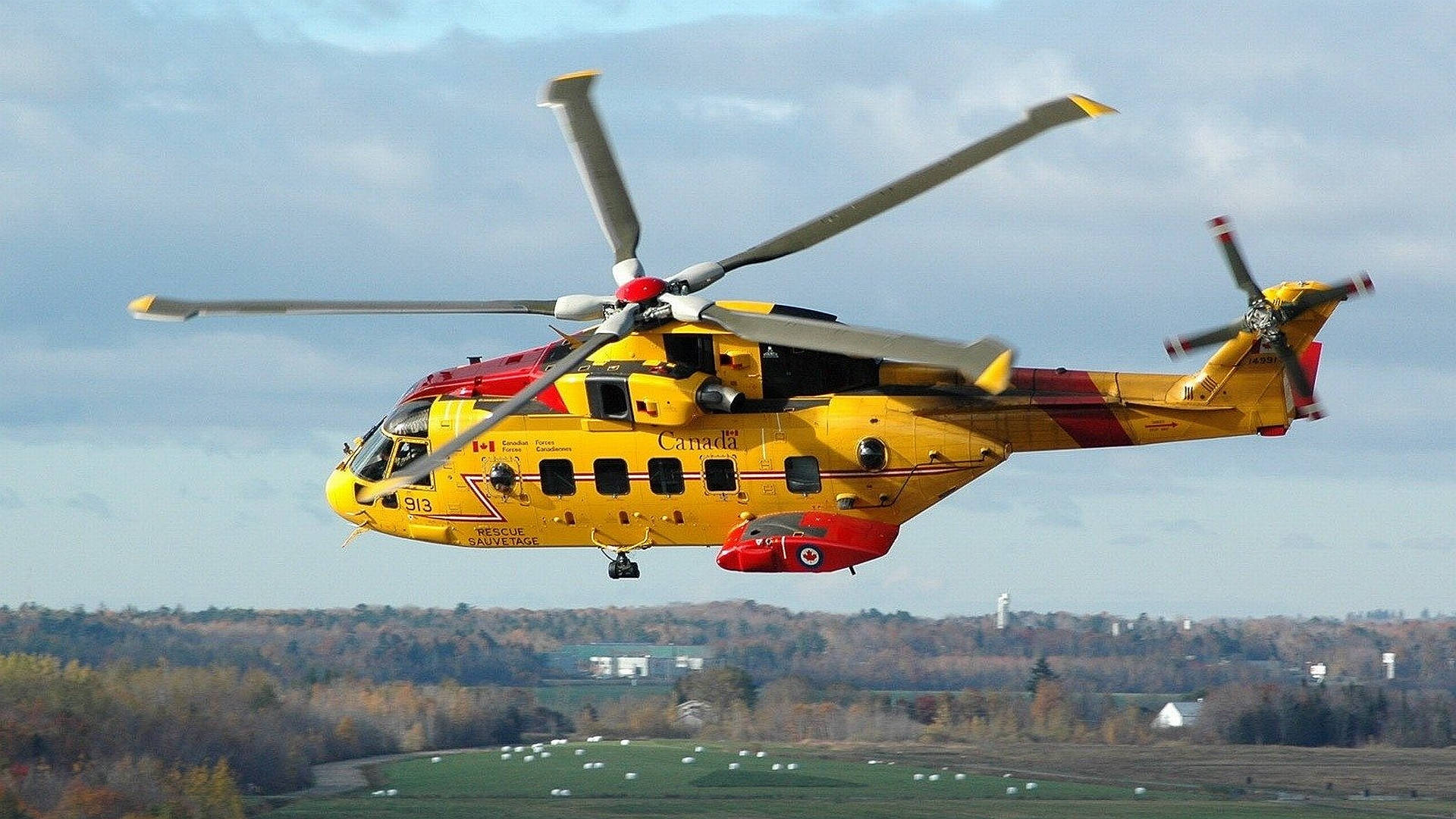 helicopter 4K 桌布 壁紙 — 一架亮黃色的加拿大搜索救援直升機在陰天低空飛過鄉村景觀，有秋天的樹木和綠色的田野。