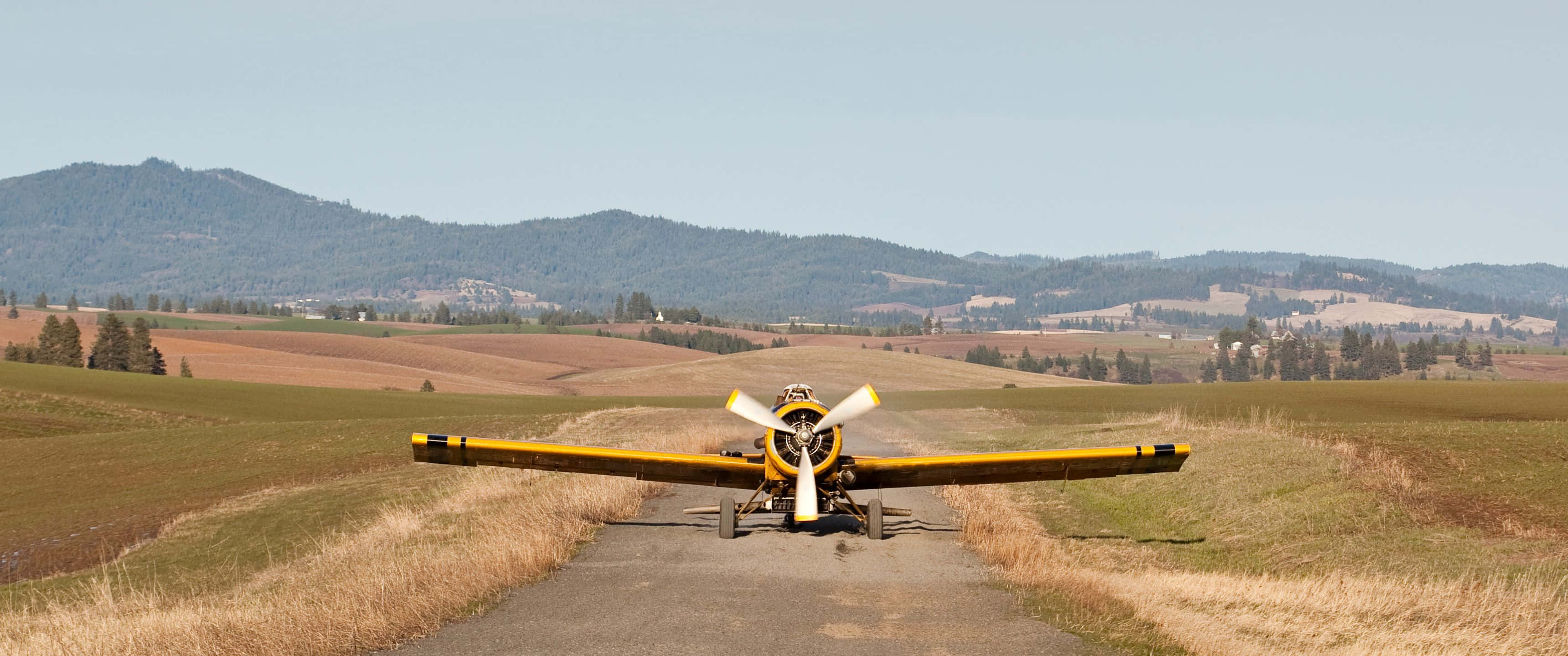 3440x1440p Small Planes 桌布 壁紙 — 一架亮黃色的復古農用噴灑飛機停泊在陽光明媚的鄉村田野的碎石路上，周圍是連綿起伏的丘陵和遠處的山脈，天空清澈湛藍。