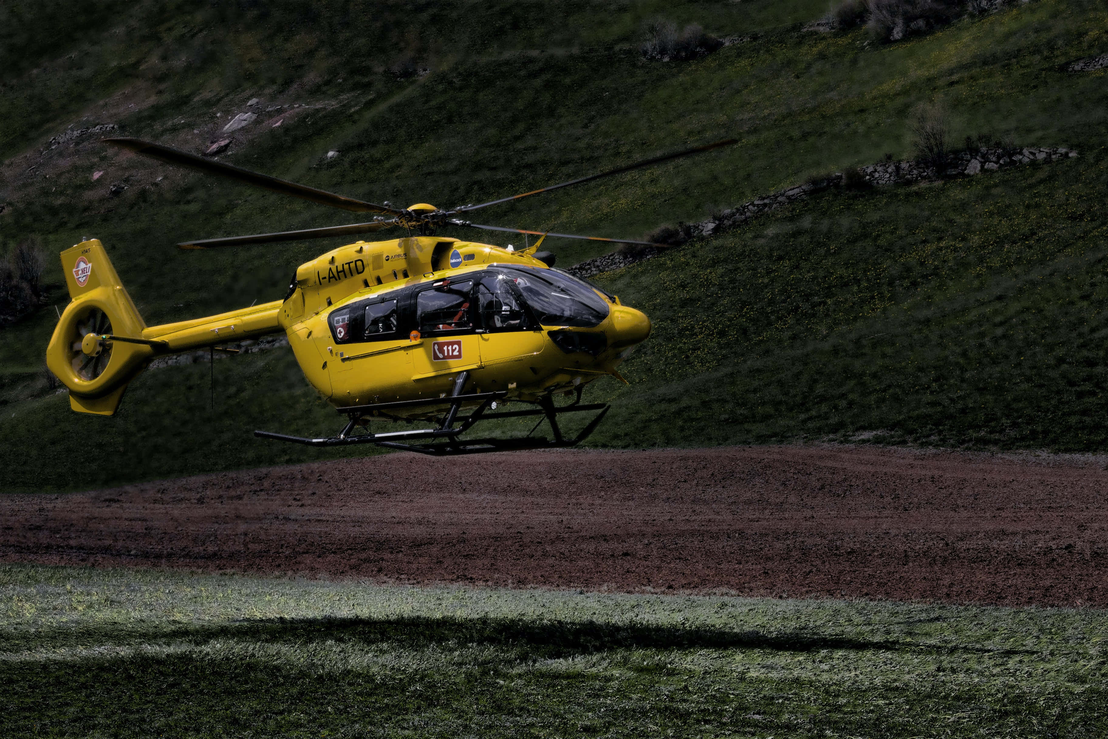 Helicopter 桌布 壁紙 — 一架亮黃色的救援直升機低空盤旋在崎嶇的山坡上，地形呈現綠色和棕色。