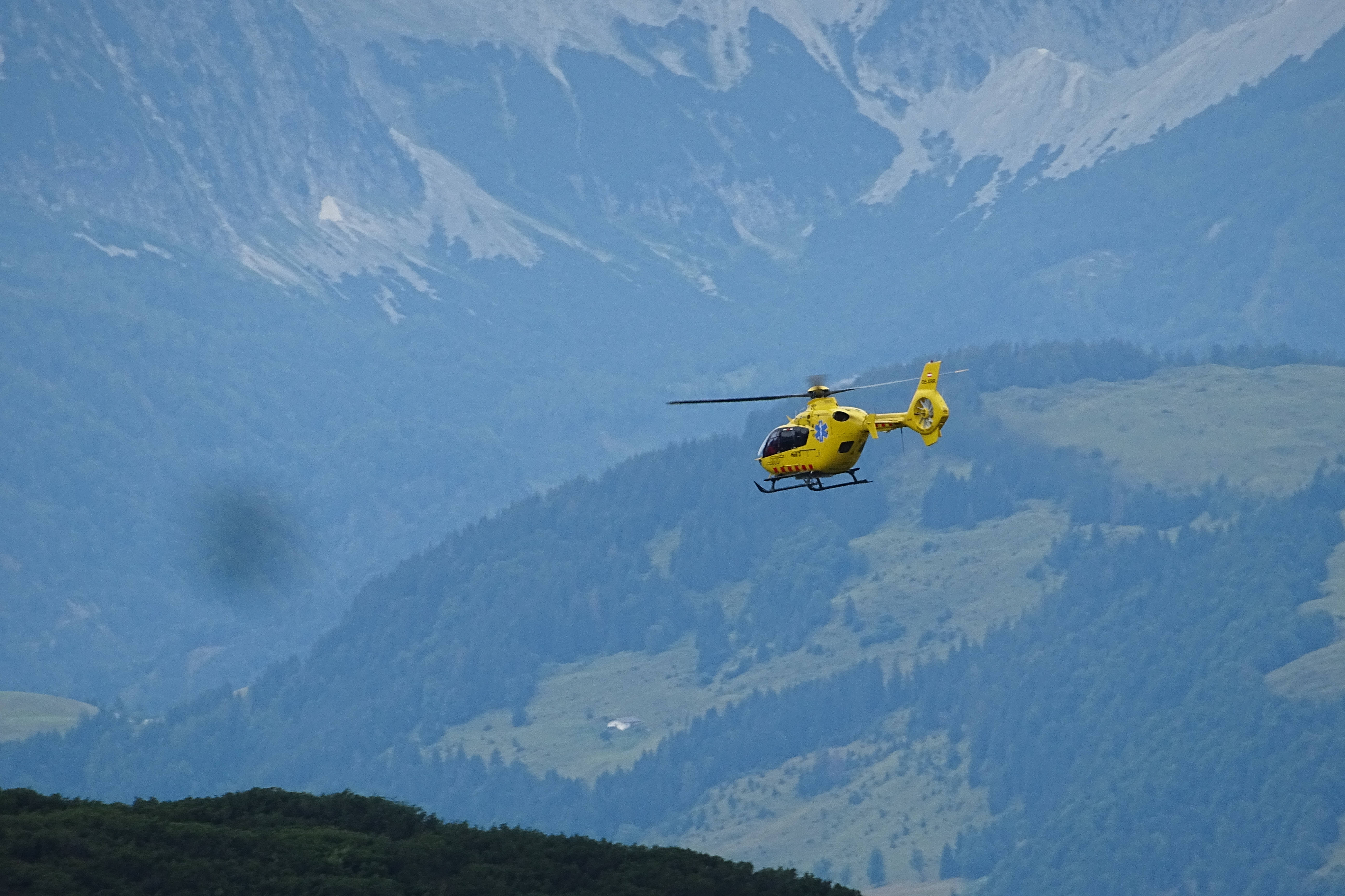 helicopter 4K 桌布 壁紙 — 一架亮黃色的救援直升機飛越雪山和森林覆蓋的山坡景觀。