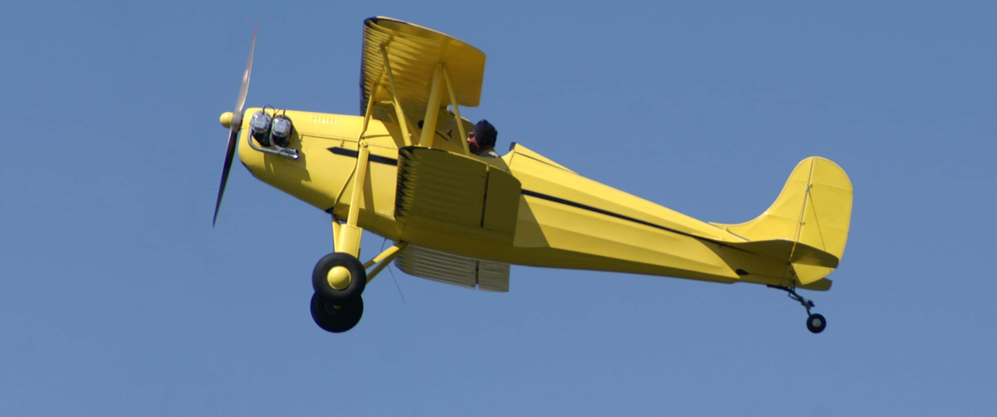 3440x1440p Small Planes 桌布 壁紙 — 一架亮黃色的雙翼飛機在清澈的藍天中翱翔，駕駛艙內可見飛行員。