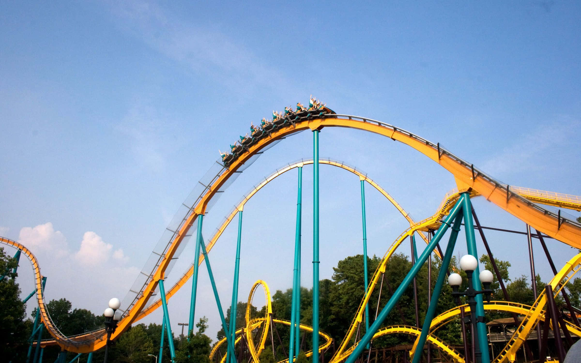 Roller Coaster 桌布 壁紙 — 一架令人興奮的黃色和藍綠色雲霄飛車在晴朗的藍天中飛馳，乘客正處於迴環頂點。