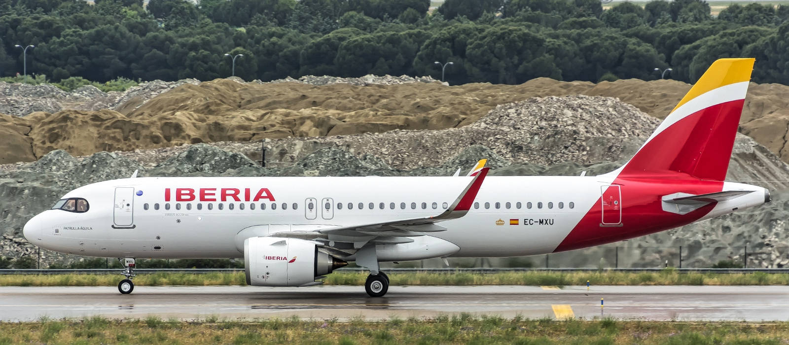 Airport 桌布 壁紙 — 一架伊比利亞航空的空中巴士A320在潮濕的跑道上滑行，背景是土色的山丘和茂密的綠樹。