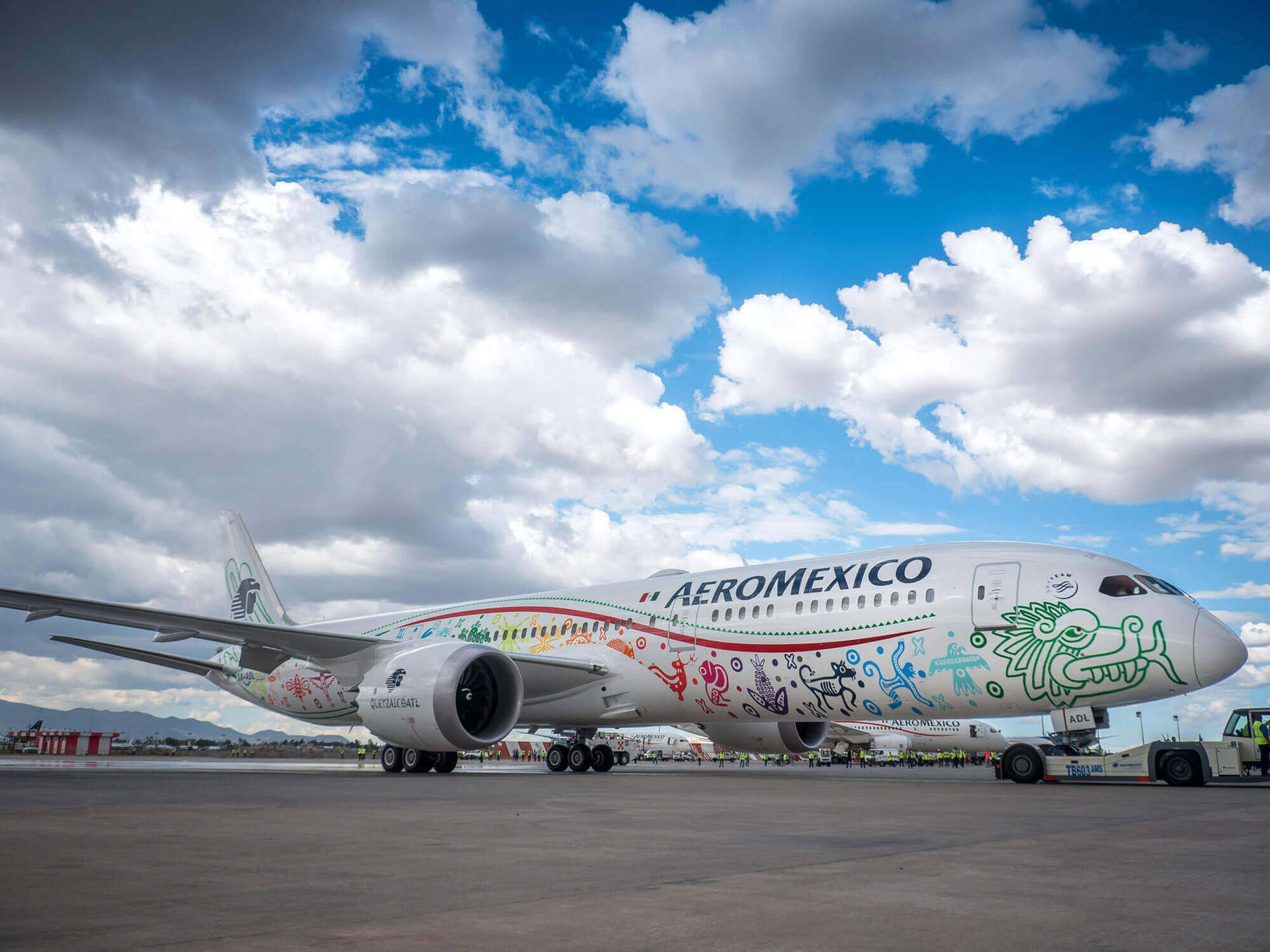 Plane 桌布 壁紙 — 一架充滿活力的墨西哥航空飛機，飾有色彩繽紛的原住民藝術，停在機場停機坪上，天空烏雲密布，氣氛戲劇化。