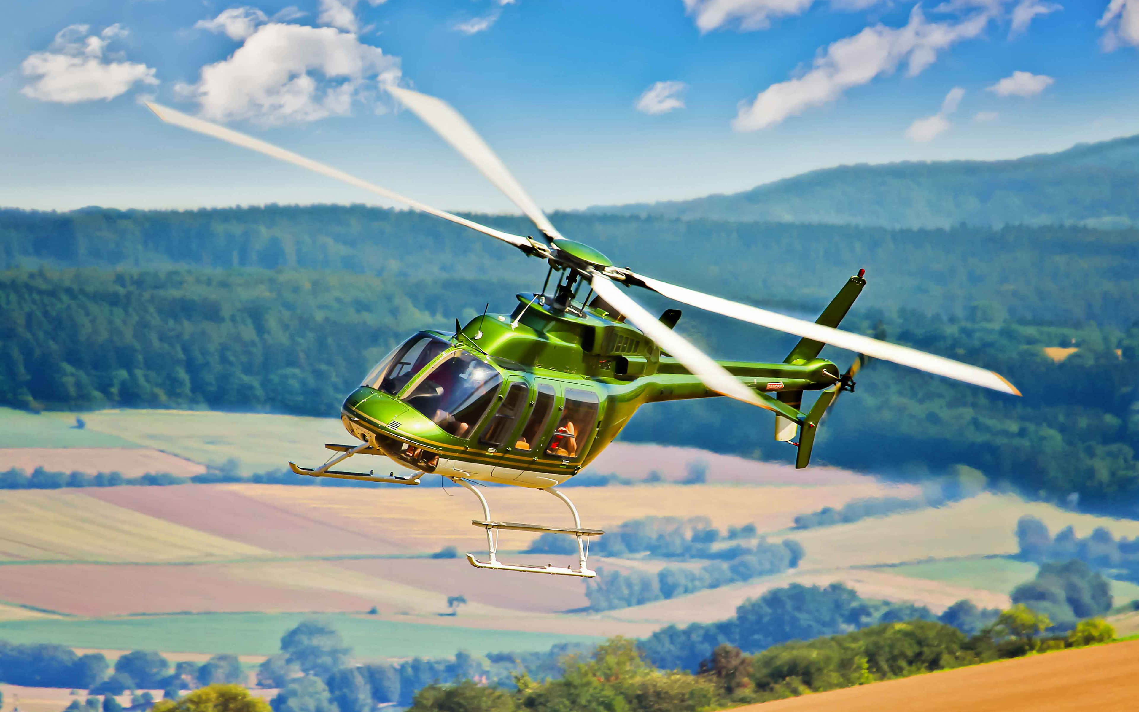 4K Helicopters 桌布 壁紙 — 一架充滿活力的綠色直升機在連綿起伏的山丘和拼布般的田野上空翱翔，背景是晴朗的藍天和蓬鬆的白雲。
