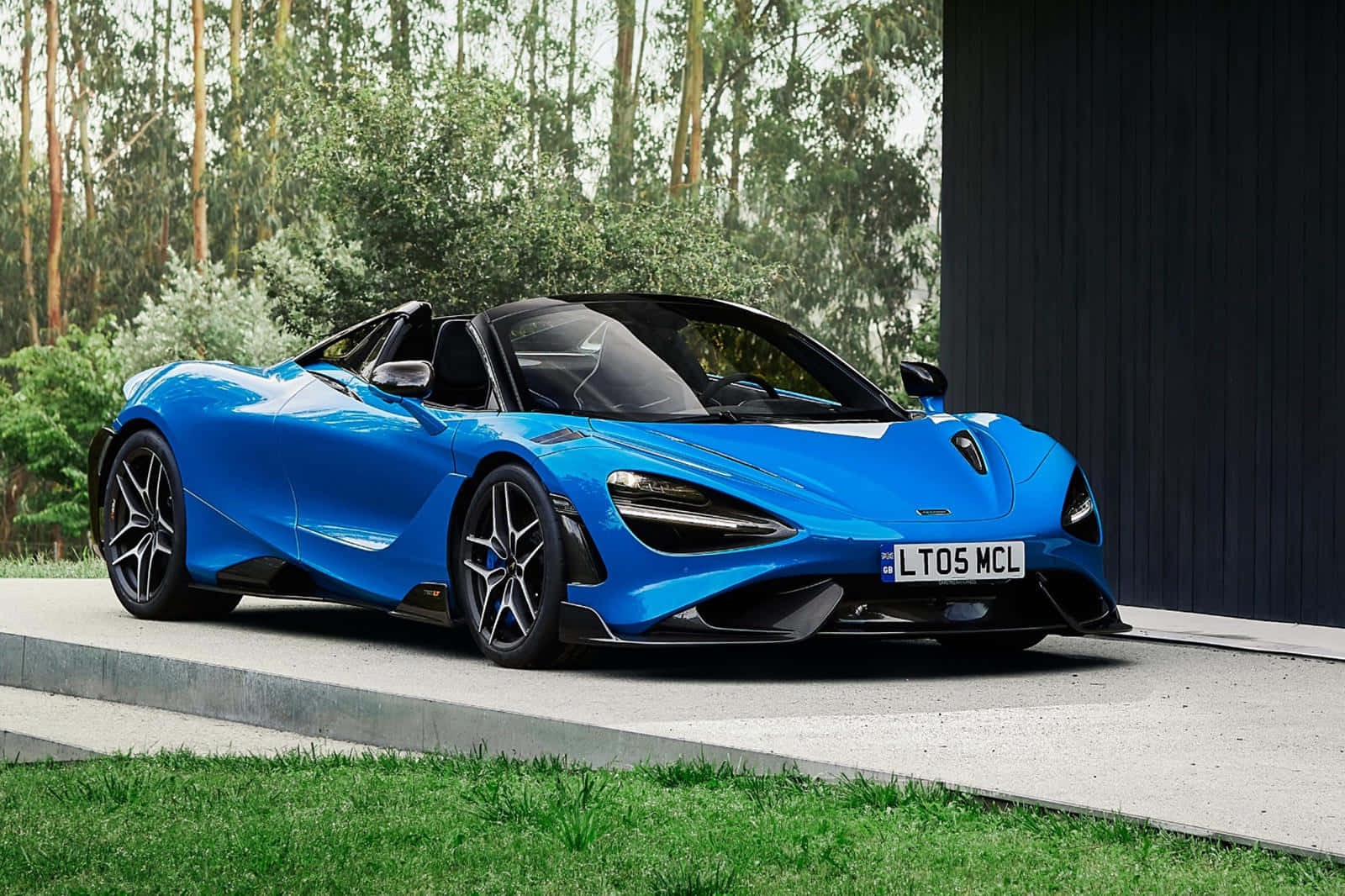 麥拉倫 桌布 壁紙 — 一架充滿活力的藍色McLaren 720S Spider敞篷跑車停放在混凝土平台上，旁邊是一座深色木建築，背景有綠樹。