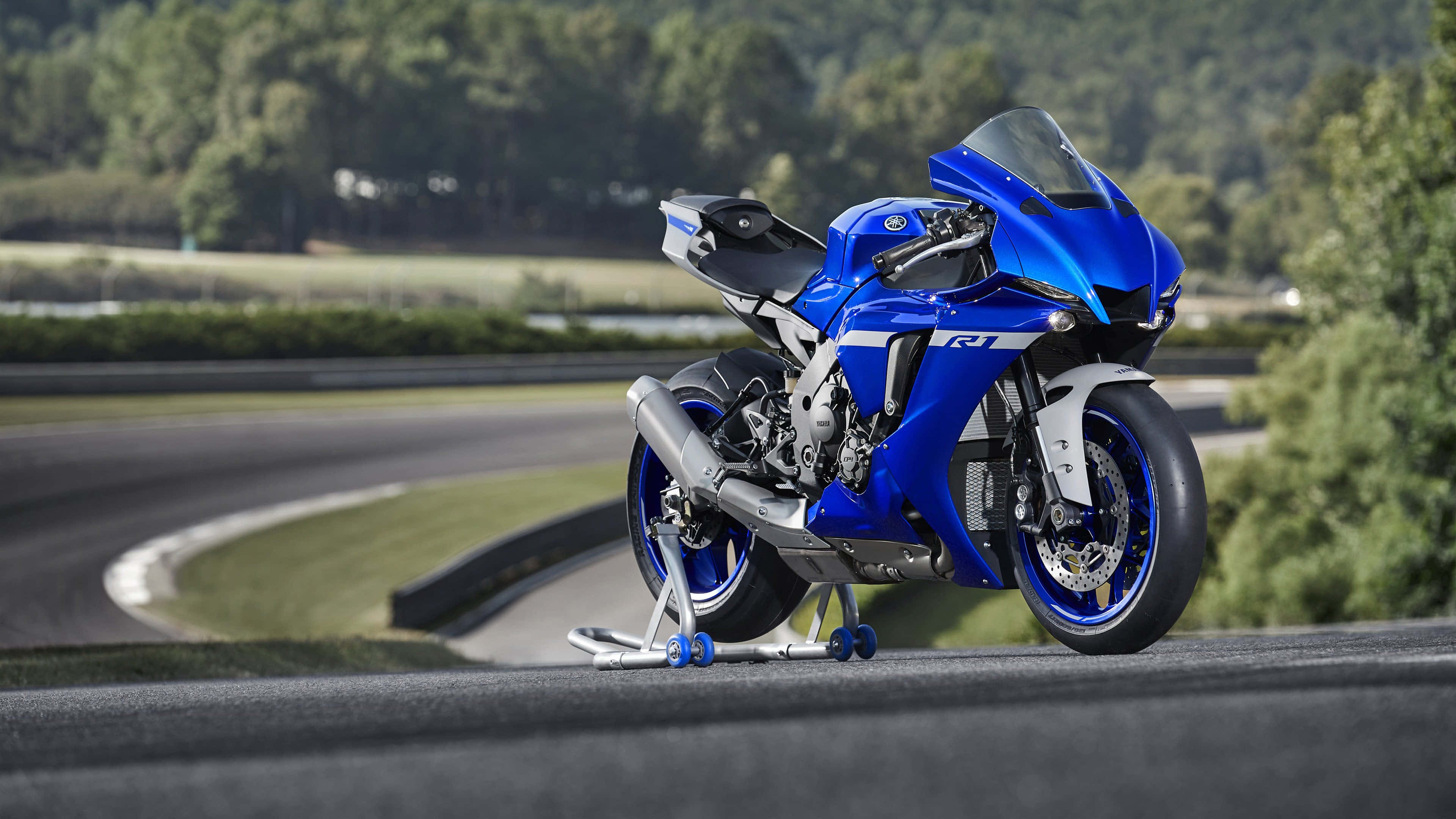 4K Bikes 桌布 壁紙 — 一架充滿活力的藍色Yamaha R1機車停在賽道上，背景是茂密的綠色樹木。