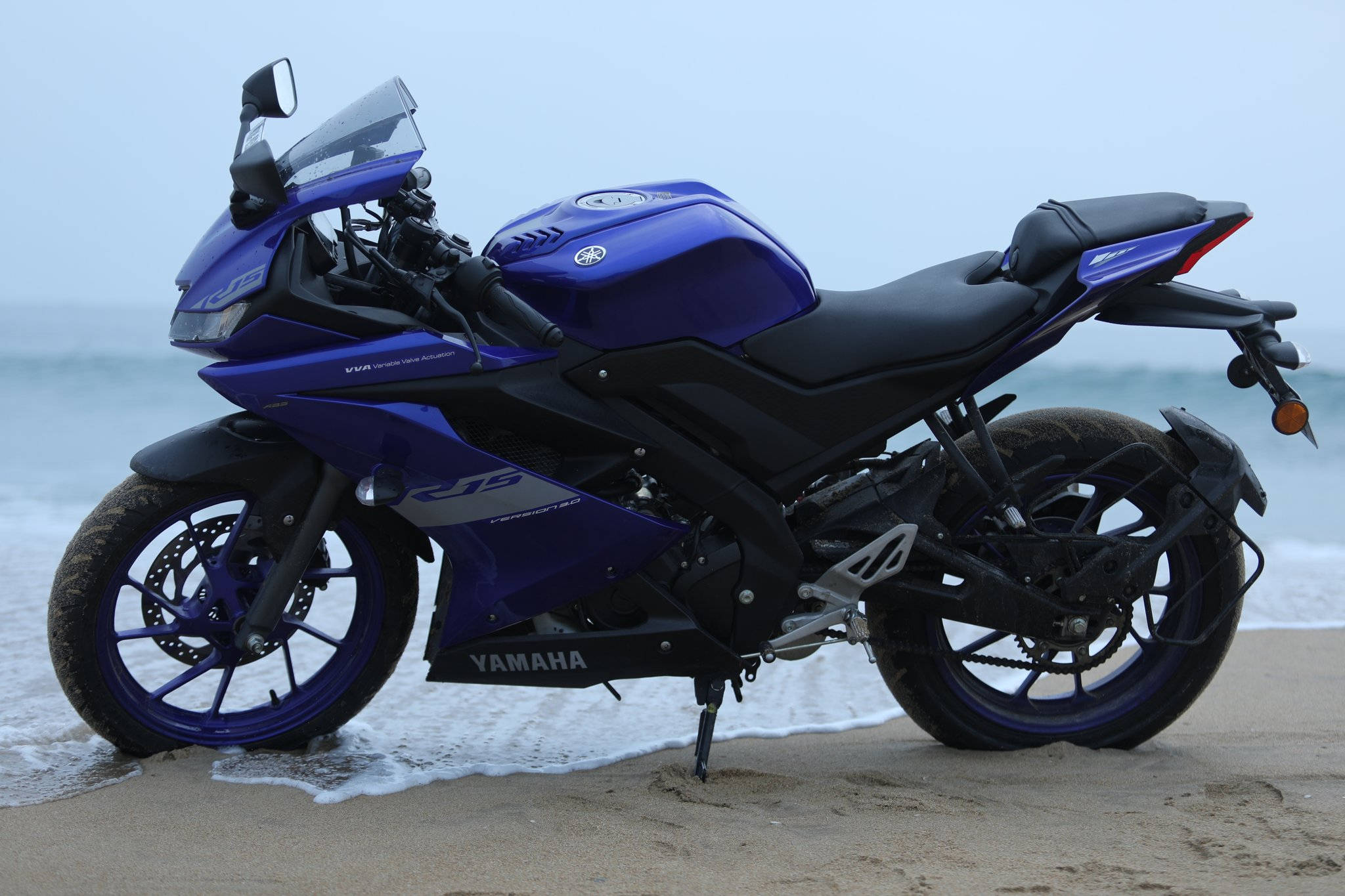桌布 壁紙 — 一架充滿活力的藍色Yamaha R15運動機車停在濕沙上，海浪在陰天輕拍其輪胎。