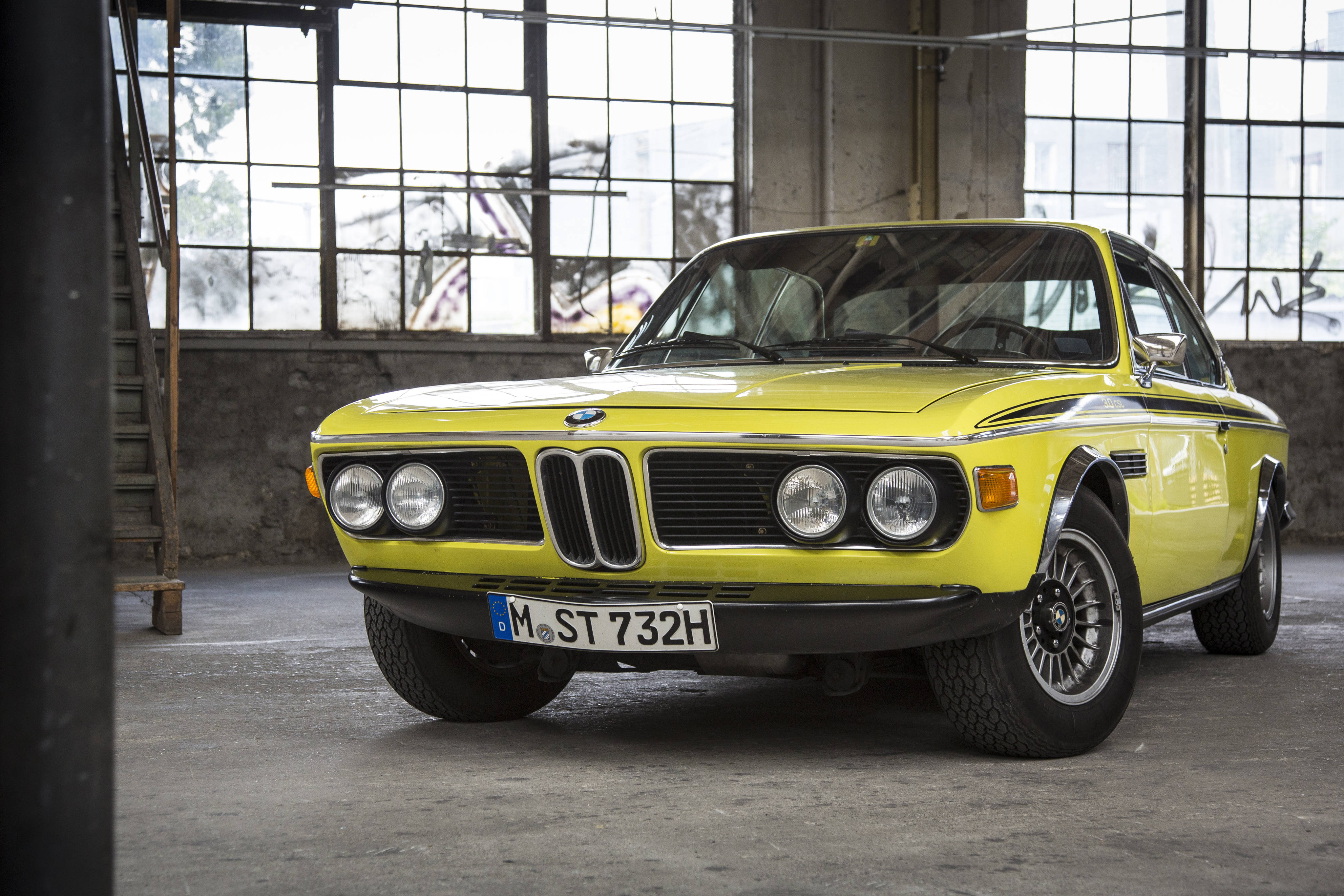 4K BMW 壁紙 桌布 壁紙 — 一架充滿活力的黃色經典BMW 2002 Turbo停放在陽光普照的工業倉庫內，倉庫有大窗戶和混凝土地板。