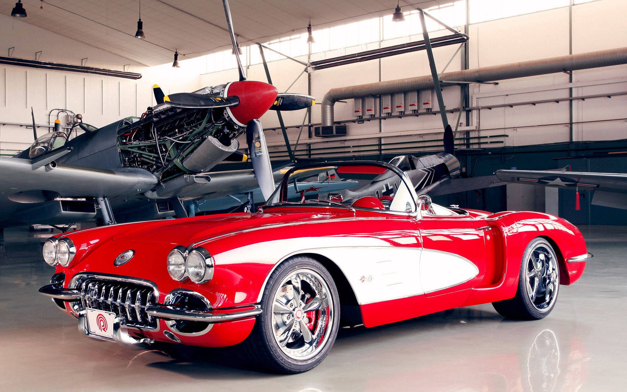 經典汽車 桌布 壁紙 — 一架光澤的紅白經典Corvette敞篷車停放在寬敞機庫內一架復古螺旋槳飛機旁邊。