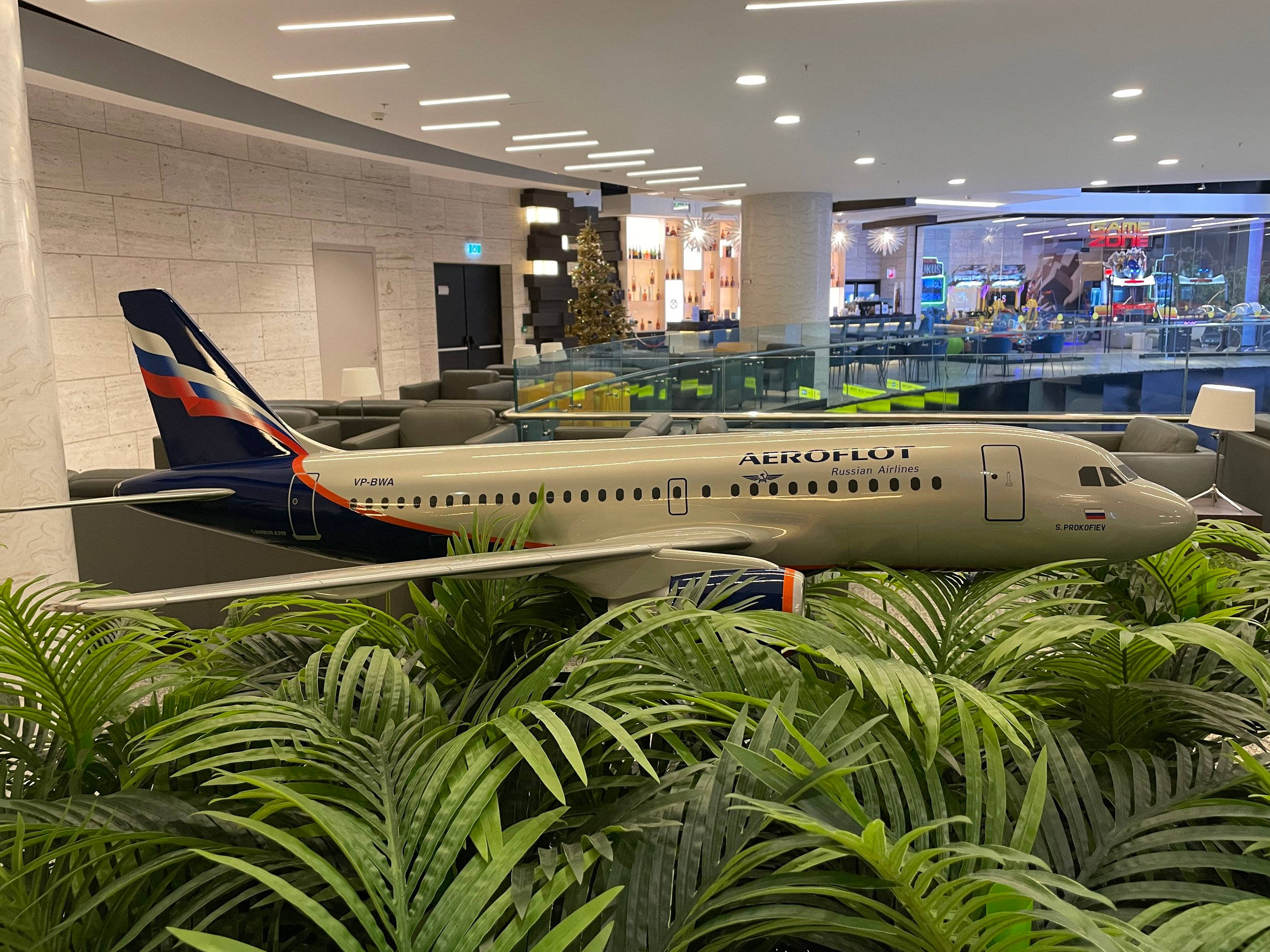 transportation 桌布 壁紙 — 一架全尺寸的Aeroflot飛機模型放置在現代機場休息室的茂密綠色蕨類植物上，配有玻璃欄杆和節日燈飾。