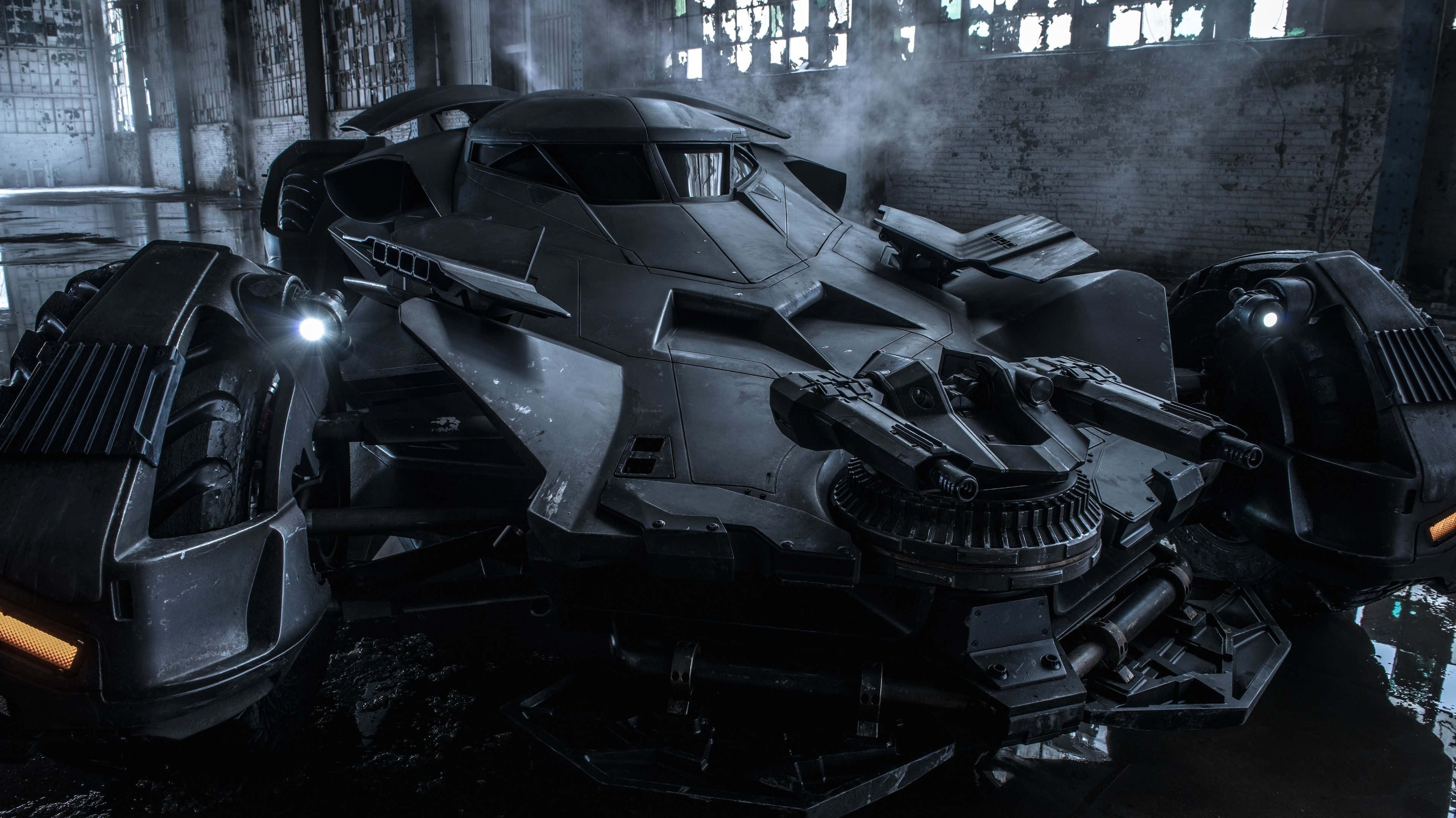 4K Batmobile 桌布 壁紙 — 一架具威脅性的黑色裝甲蝙蝠車停放在一個被水淹沒、煙霧瀰漫、窗戶破爛的廢棄倉庫中。